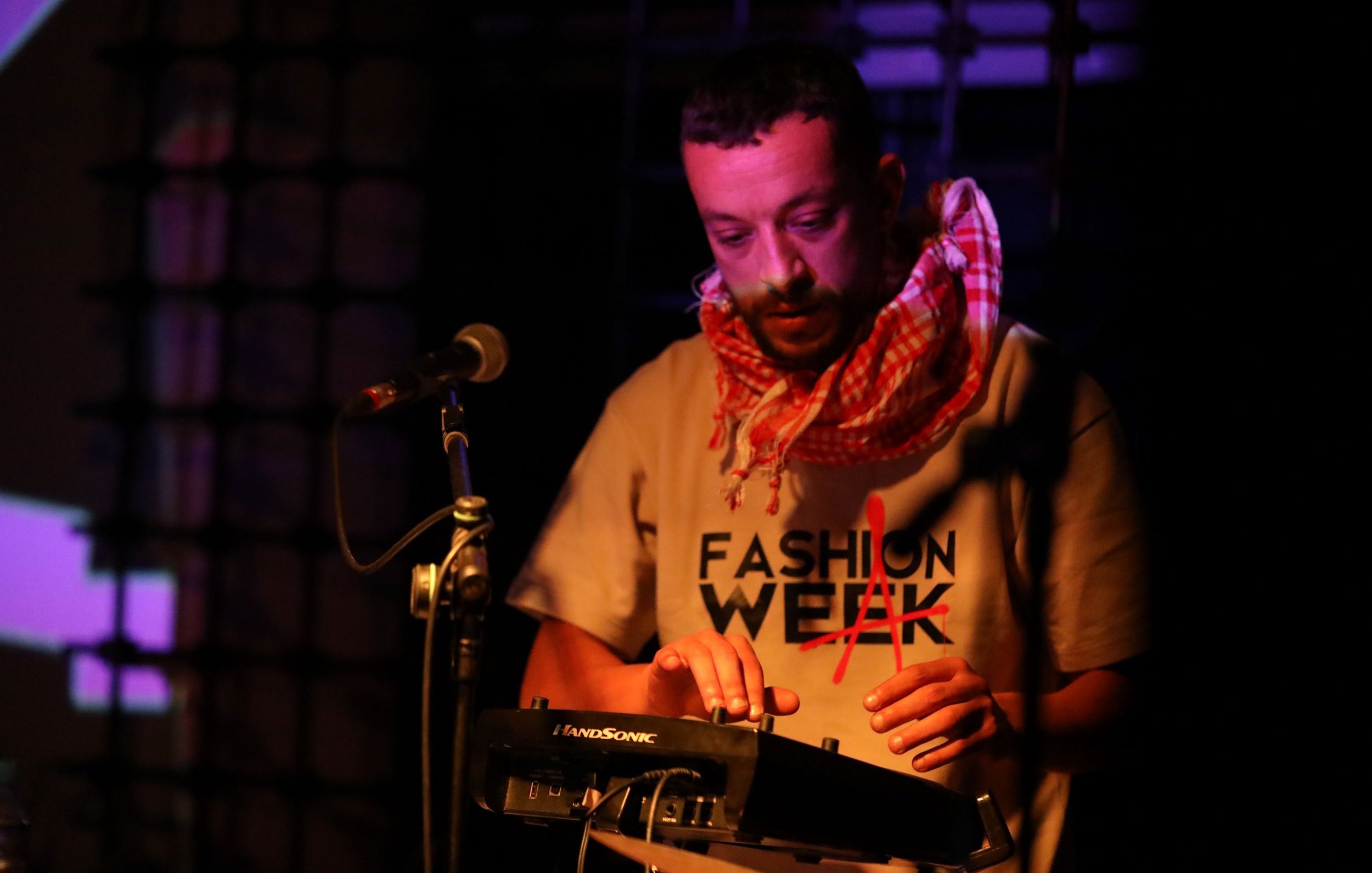 L'image montre un homme en train de performer sur scène. Il porte un t-shirt avec le mot "FASHION" écrit dessus et un foulard autour du cou. Il utilise un petit appareil électronique, probablement un synthétiseur ou un contrôleur musical. Le fond est légèrement éclairé avec des lumières colorées, créant une ambiance dynamique. Le microphone est également visible à côté de lui, indiquant qu'il pourrait chanter ou parler en même temps. L'atmosphère semble électrique et captivante.