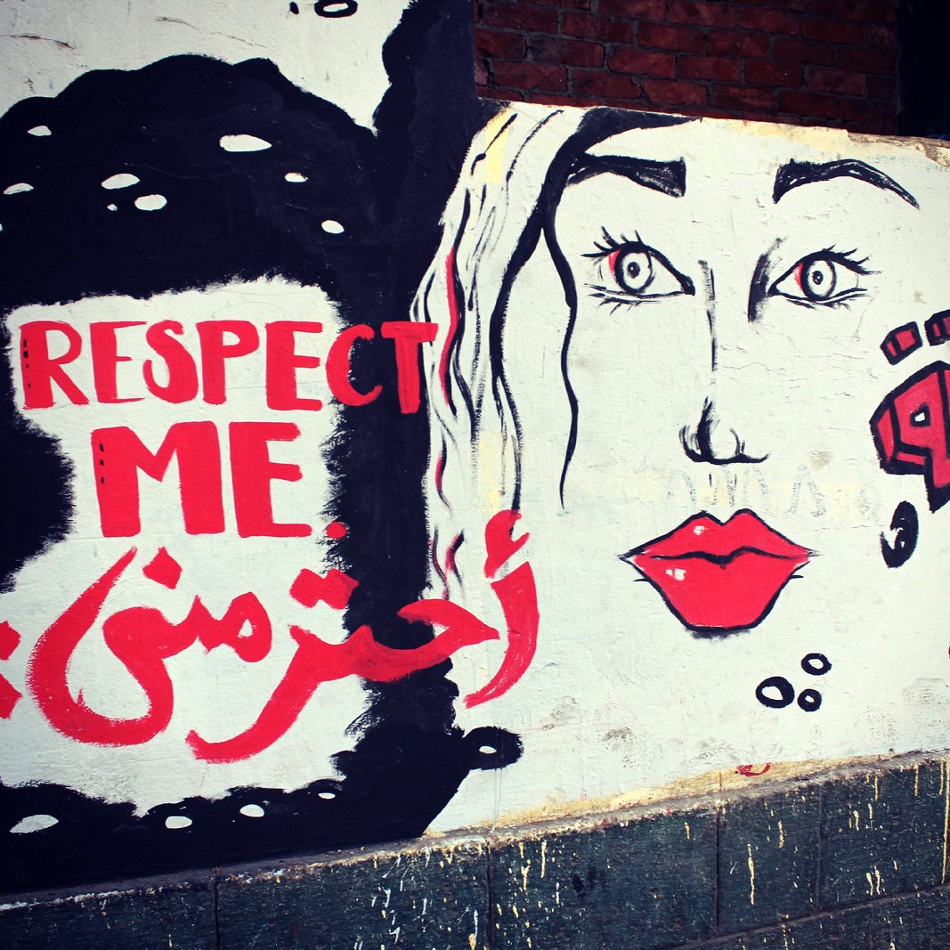 L'image montre une fresque murale qui combine des éléments graphiques et des mots en anglais et en arabe. À gauche, on peut voir le texte "RESPECT ME" en anglais, et en dessous, "احترمني" qui signifie "Respecte-moi" en arabe. À droite, il y a un visage stylisé de femme, avec des yeux expressifs et des lèvres rouges. Le tout est peint sur un mur de couleur claire, créant un message fort sur l'importance du respect.