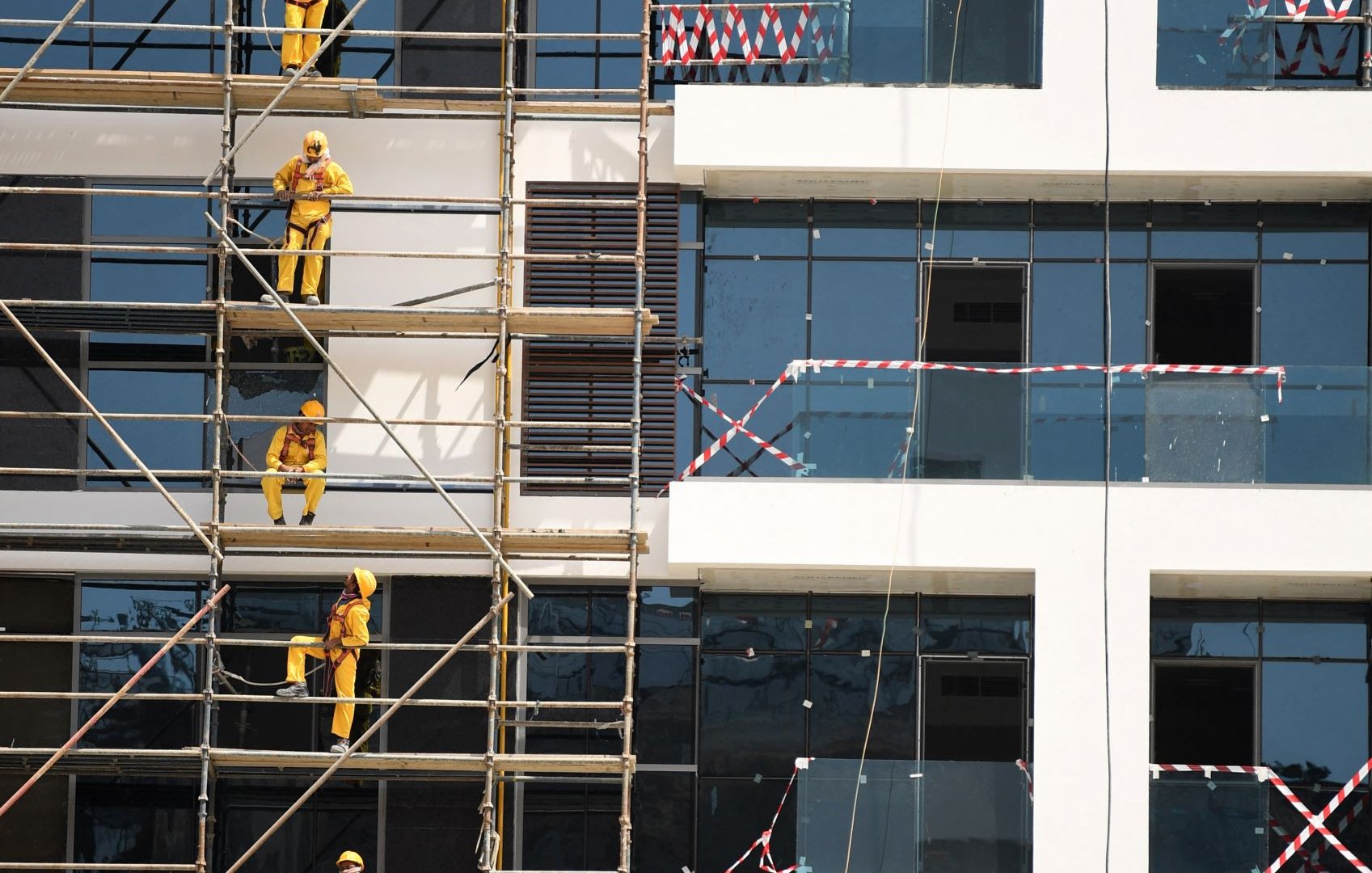L'image montre un bâtiment en construction. On peut voir des échafaudages le long de la façade, sur lesquels plusieurs ouvriers vêtus de combinaisons jaunes et de casques de sécurité sont en train de travailler. Certains utilisent des outils pour peindre ou poser des éléments sur les fenêtres, tandis que d'autres montent et descendent sur les échafaudages. Le bâtiment a des fenêtres en verre, et des bandes de sécurité sont visibles autour de certaines zones. L'ensemble de la scène reflète un environnement de travail actif et organisé.