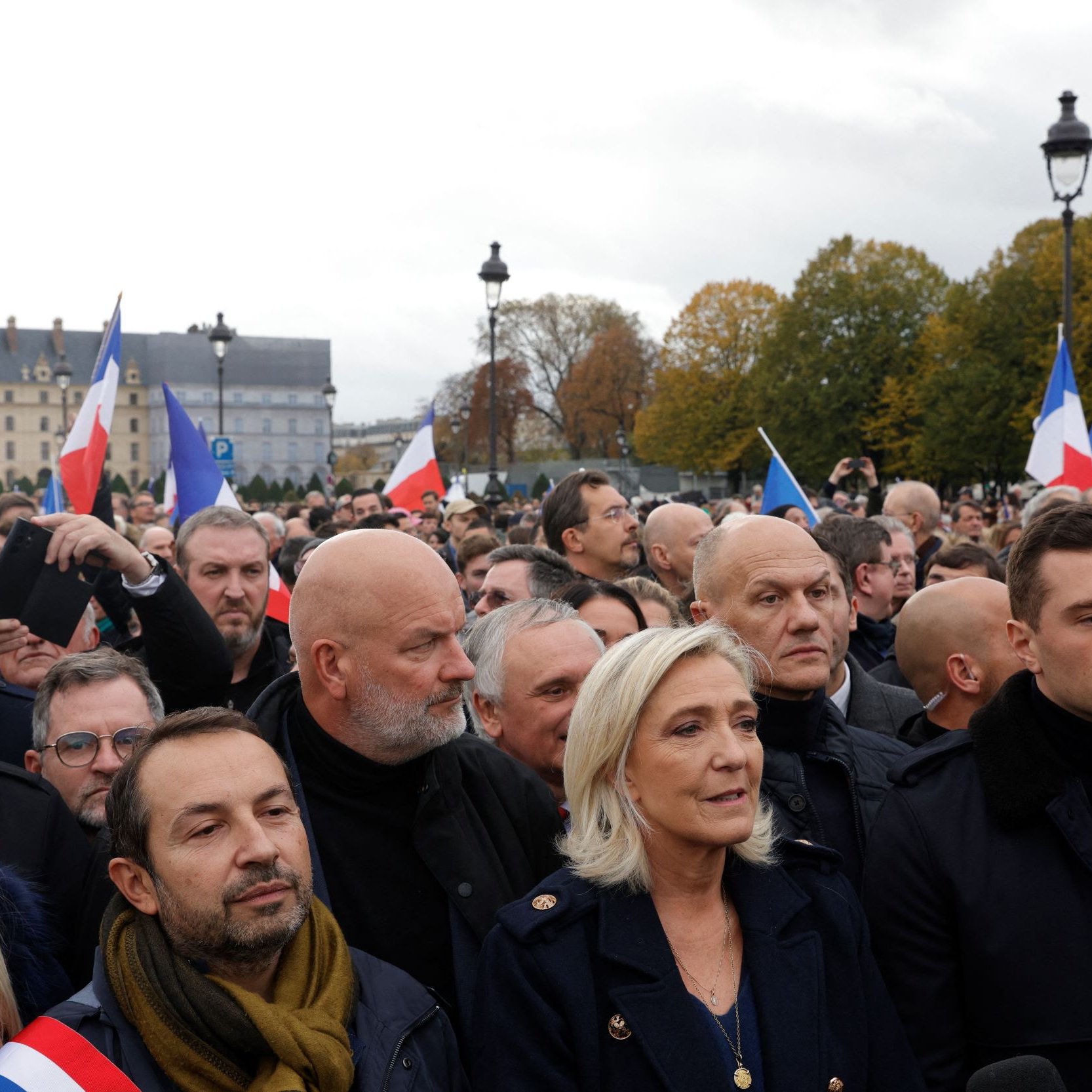 The image depicts a crowd of people gathered in an outdoor setting, likely at a political event or rally. Many individuals are holding French flags, and the expressions on their faces suggest a mix of determination and focus. Prominent figures can be seen at the forefront, wearing formal attire. The background features historic architecture and trees, indicating an urban environment. The atmosphere appears charged, reflecting the significance of the gathering.