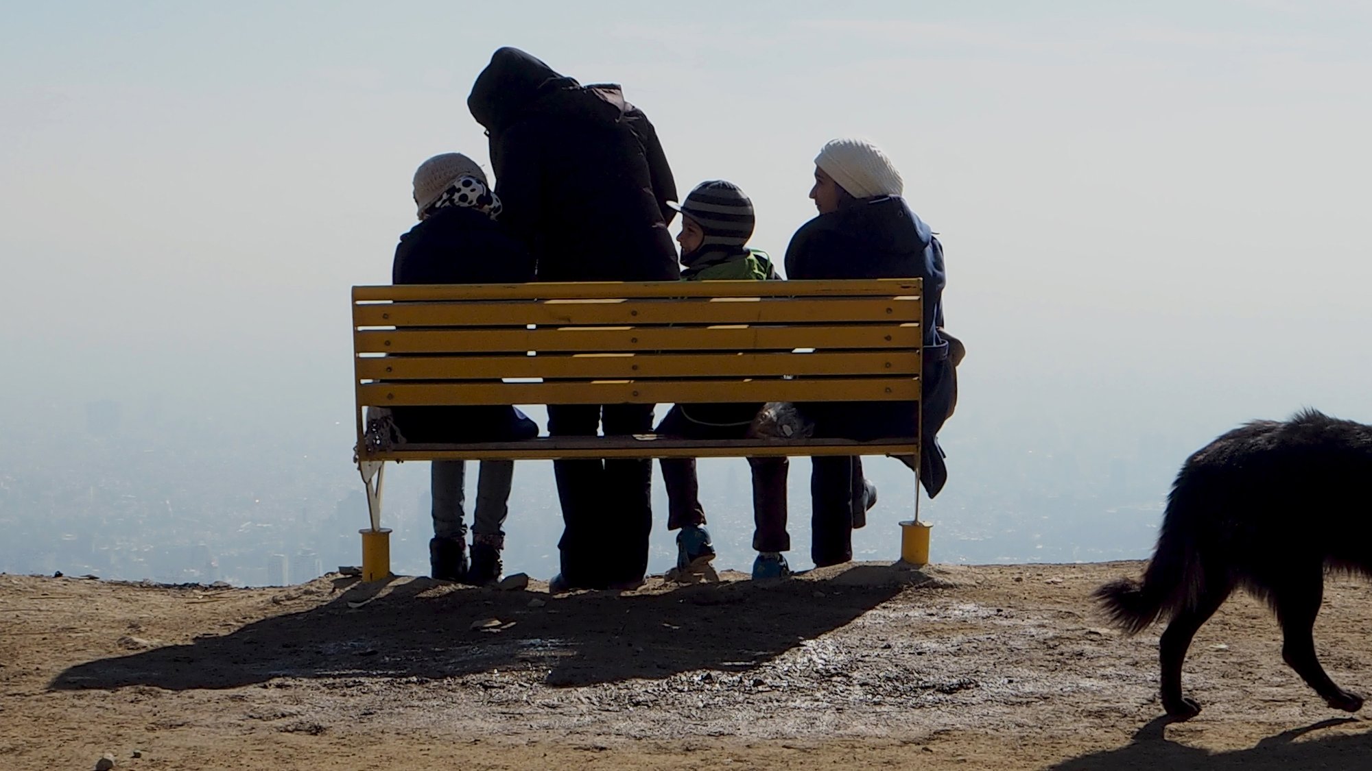 در این تصویر، تعدادی از افراد در حال نشستن بر روی یک نیمکت زرد رنگ هستند. آنها به سمت یک چشمانداز زیبا و دوردست نگاه میکنند. چهار نفر در صحنه دیده میشوند: سه نفر از آنها نشستهاند و یک نفر، که احتمالاً بزرگتر است، پشت به دوربین ایستاده و به آنها توجه دارد. هوا صاف به نظر میرسد و درای پسزمینهای مهآلود است. همچنین یک سگ سیاه رنگ نیز در گوشه تصویر دیده میشود. حس کلی تصویر آرامش و دوستی را منتقل میکند.