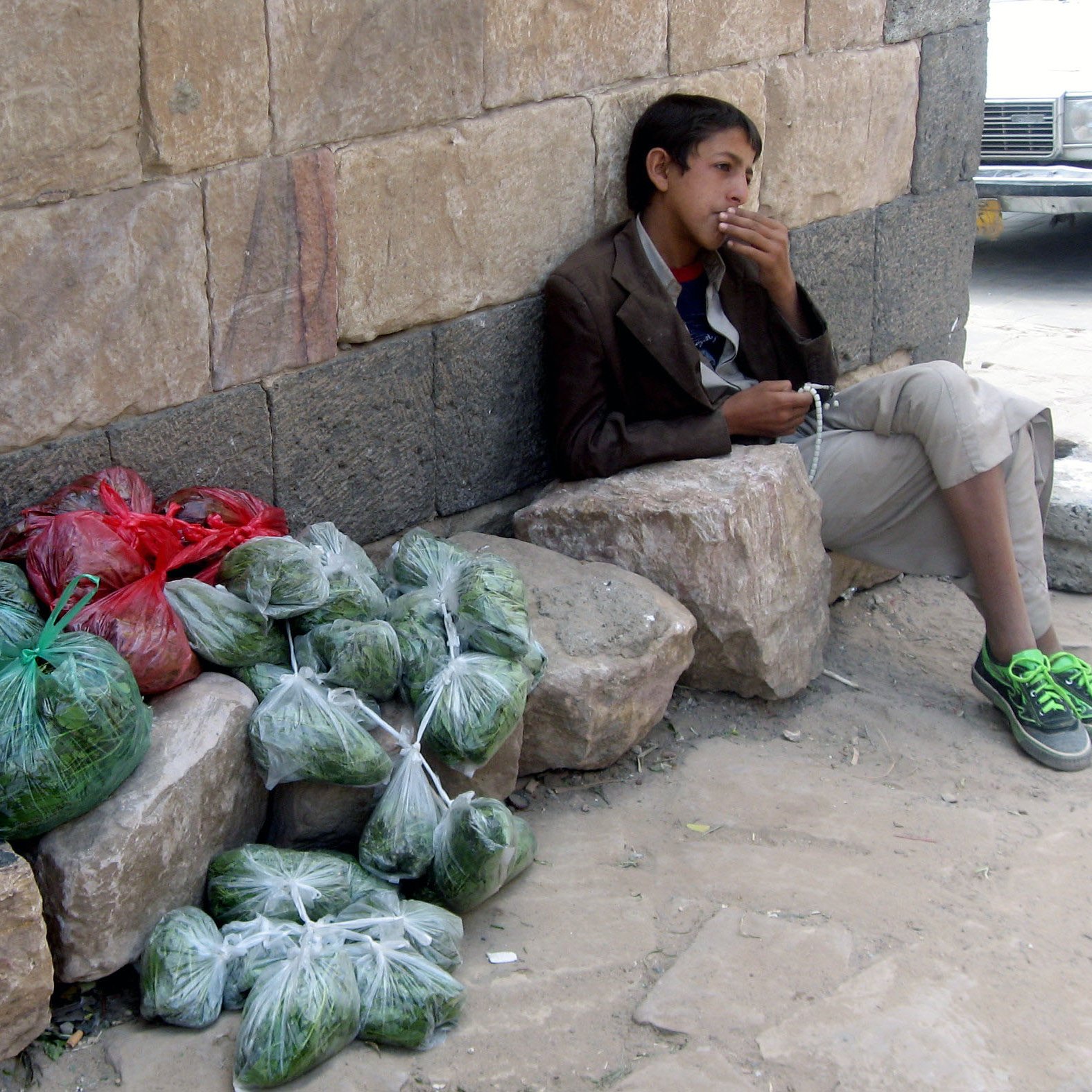 في الصورة، يظهر فتى جالساً بجوار حائط حجري. يتكئ على صخرة كبيرة وينظر إلى المسافة. يرتدي ملابس بسيطة، ويبدو أنه في حالة تفكير أو انتظار. بالقرب منه، توجد أكياس ملونة تحتوي على خضروات، بعضها بالأخضر والآخر باللون الأحمر، موضوعة بشكل منظم على الأرض. تعكس الصورة أجواء حياة الشارع وبساطة الحياة اليومية.