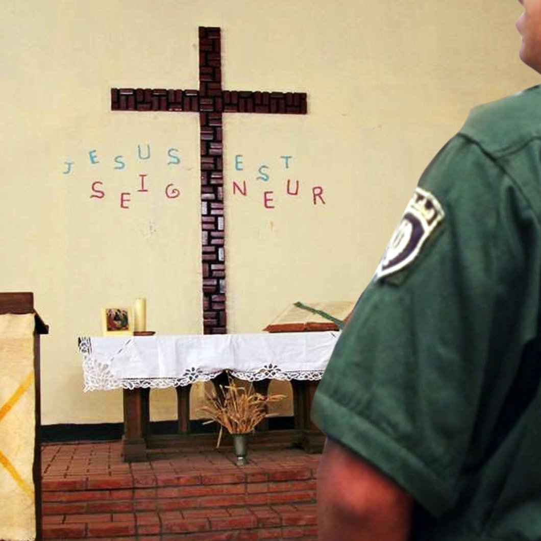 تظهر الصورة مشهداً داخل كنيسة، حيث يتواجد صليب خشبي كبير على الحائط. خلف الصليب، توجد كلمات مكتوبة باللغة الفرنسية تقول "يسوع هو الرب" (Jésus est Seigneur). في الجهة اليمنى من الصورة، يقف رجل يرتدي زيًا رسميًا ويرتدي شعارًا يدل على "الدرك الوطني". يبدو أن الرجل يراقب المكان حوله. الأجواء تعكس مزيجًا من الروحانية والتواجد الأمني.