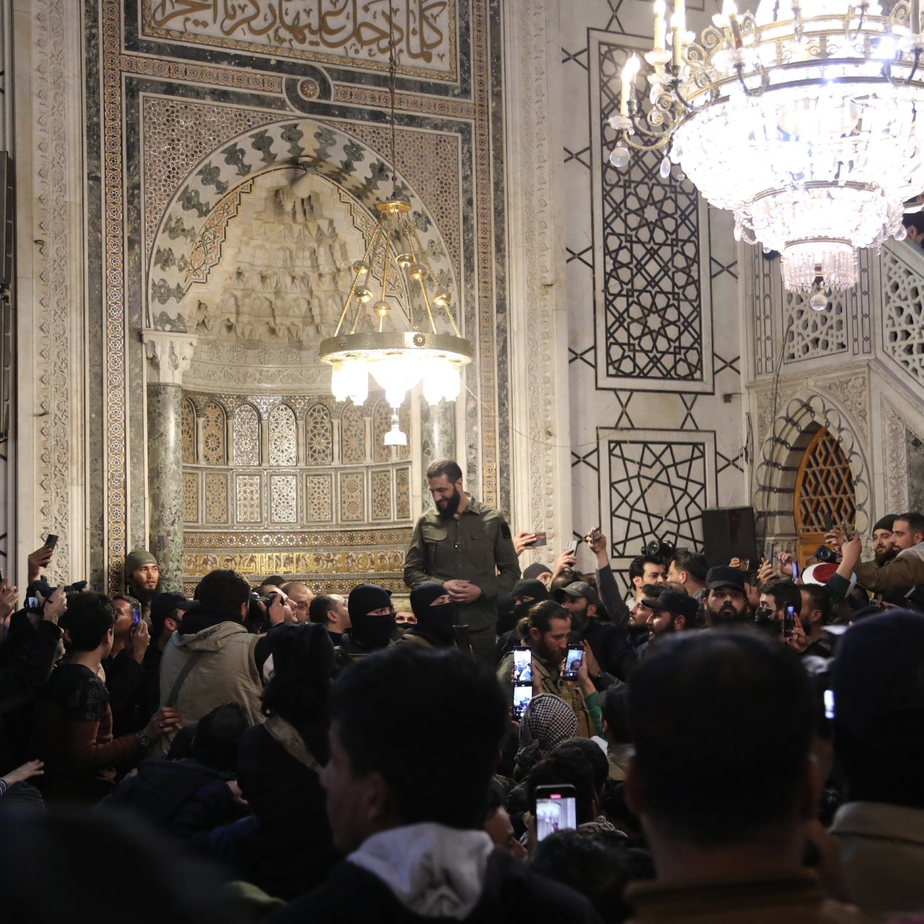 L'image montre un intérieur richement décoré, probablement d'un bâtiment historique ou religieux, avec des motifs géométriques et des ornements élaborés. Au centre, une personne se tient en hauteur, entourée d'une foule qui utilise des téléphones pour capturer le moment. Des lumières brillantes, notamment un lustre, illuminent la scène. L'atmosphère semble être celle d'un rassemblement ou d'un événement significatif.