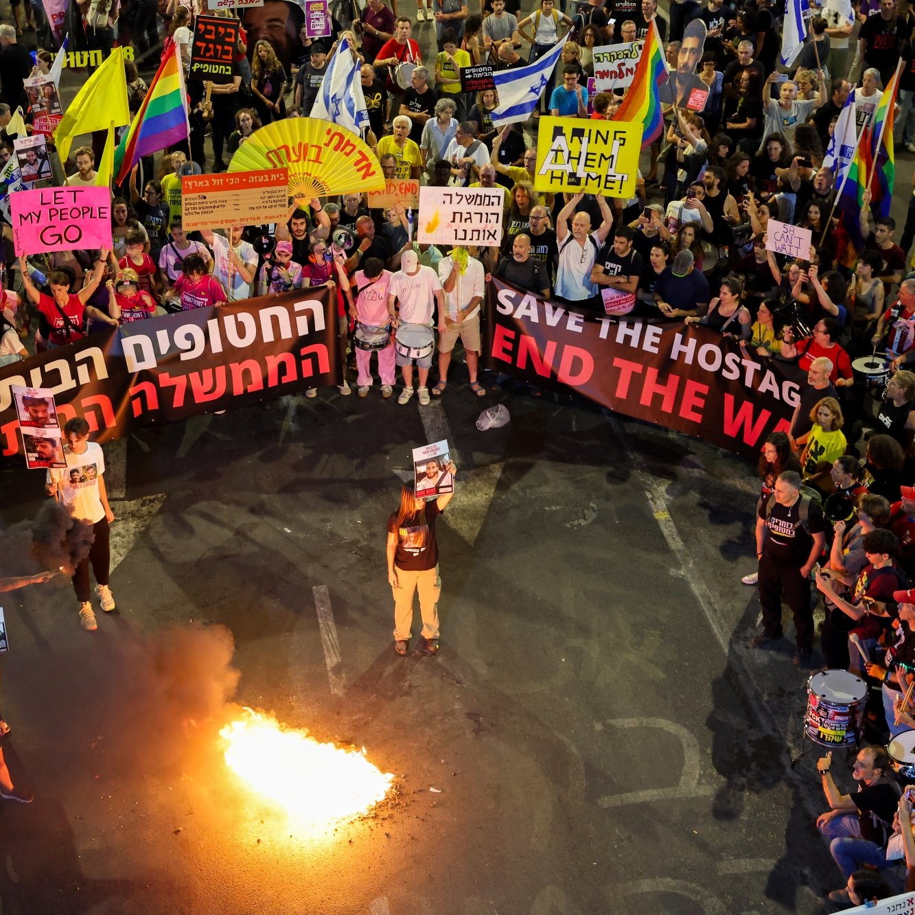 A large protest with colorful banners, people holding signs, and a fire in the center.