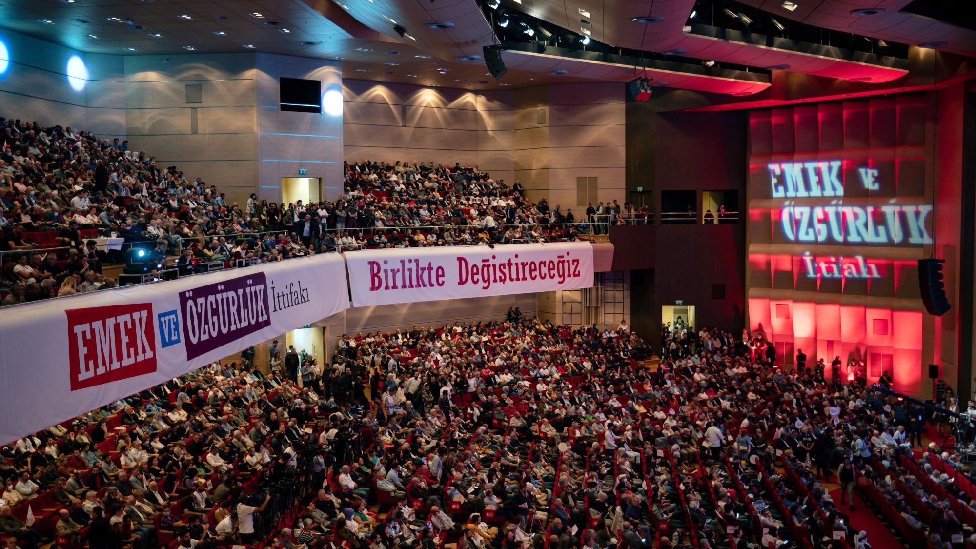 L'image montre une grande salle de conférence ou de réunion, remplie de personnes assises qui semblent assister à un événement. Les spectateurs sont disposés sur des sièges rouges, et des banderoles avec des inscriptions en turc sont accrochées sur les murs, indiquant probablement des slogans liés à des thèmes de travail ou de liberté. Un éclairage rouge illumine le fond de la scène, où des mots supplémentaires sont projetés, renforçant l'atmosphère d'un rassemblement important.
