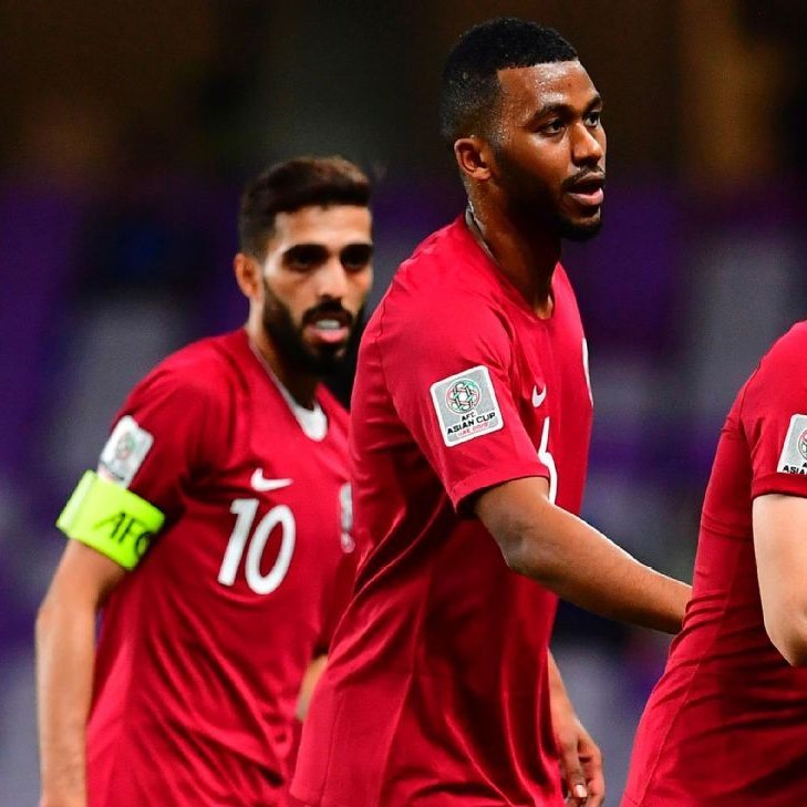 The image captures a moment from a football (soccer) match featuring players in red jerseys representing Qatar. One player, wearing the number 15, is celebrating and pulling his jersey up, possibly revealing a symbol or crest underneath. The player in the foreground seems excited and is likely celebrating a goal. Behind him, two other teammates are visible; one is wearing the number 10 and the other appears to be the team captain, as indicated by the captain's armband on his arm. The stadium setting suggests a significant match, likely during a tournament.