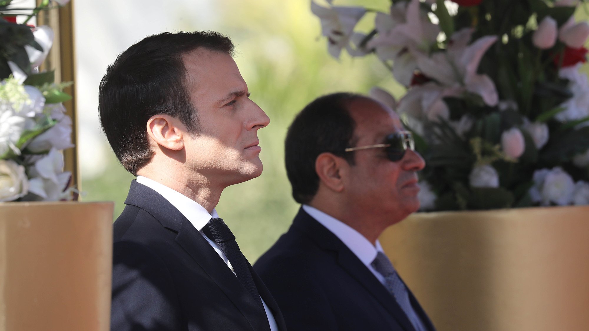 الصورة تظهر رجلين standing بجانب بعضهما البعض. الرجل على اليسار يرتدي بدلة داكنة مع ربطة عنق، بينما الرجل على اليمين يرتدي نظارات شمسية وبدلة رسمية. الخلفية تحتوي على زهور وأشجار، مما يضفي جوًا من الاحتفال أو المناسبة الرسمية. تعكس الصورة موقفًا رسميًا وجديًا بين الشخصين.