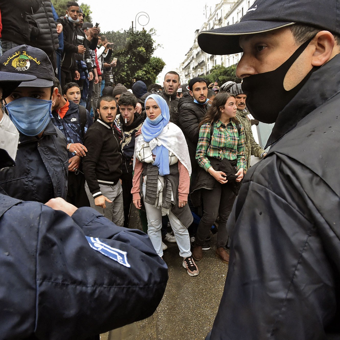 L'image montre une scène de tension entre des personnes en masse et des policiers. On peut apercevoir plusieurs individus, dont une femme portant un hijab et des vêtements clairs, qui semblent interagir avec les forces de l'ordre. Les policiers, en uniforme et portant des masques, sont en position d'encadrer la situation. L'arrière-plan montre une foule, indiquant un rassemblement ou une manifestation, et le temps semble pluvieux, ce qui ajoute à l'atmosphère de la scène.