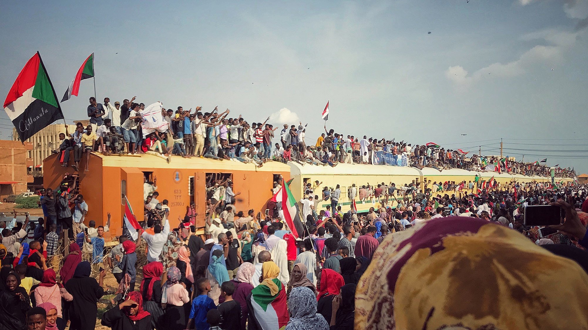 L'image montre une grande foule de manifestants rassemblés autour d'un train. De nombreuses personnes sont sur le toit et les côtés du train, brandissant des drapeaux, notamment des drapeaux soudanais. L'atmosphère semble énergique et festive, avec des gens qui expriment leur enthousiasme et leur solidarité. Le ciel est partiellement nuageux, et la scène donne une impression de mouvement et d'engagement collectif.