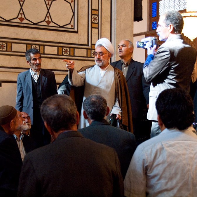 L'image montre un groupe de personnes rassemblées dans un espace intérieur richement décoré. Au centre, un homme en vêtements traditionnels, probablement religieux, semble s'adresser à l'assemblée. Il lève une main en signe d'argumentation ou d'expression. Plusieurs autres hommes l'écoutent attentivement, tandis qu'une ou deux femmes en arrière-plan portent un hijab. L'atmosphère semble sérieuse et respectueuse, avec des détails architecturaux élégants autour.