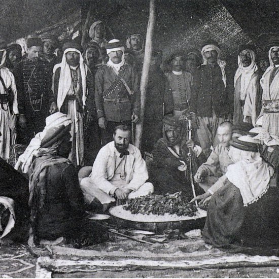 The image depicts a historical gathering of people, likely in a desert or rural setting. In the center, a group of men is seated around a large circular dish, which appears to contain food. They are dressed in traditional attire, with some wearing turbans and others in military or colonial-style uniforms. The background features a tent or canopy, suggesting a communal or ceremonial atmosphere. The mood seems convivial, as the participants engage with one another, possibly sharing a meal or discussing important matters. Overall, the scene reflects cultural practices and social interactions from the past.