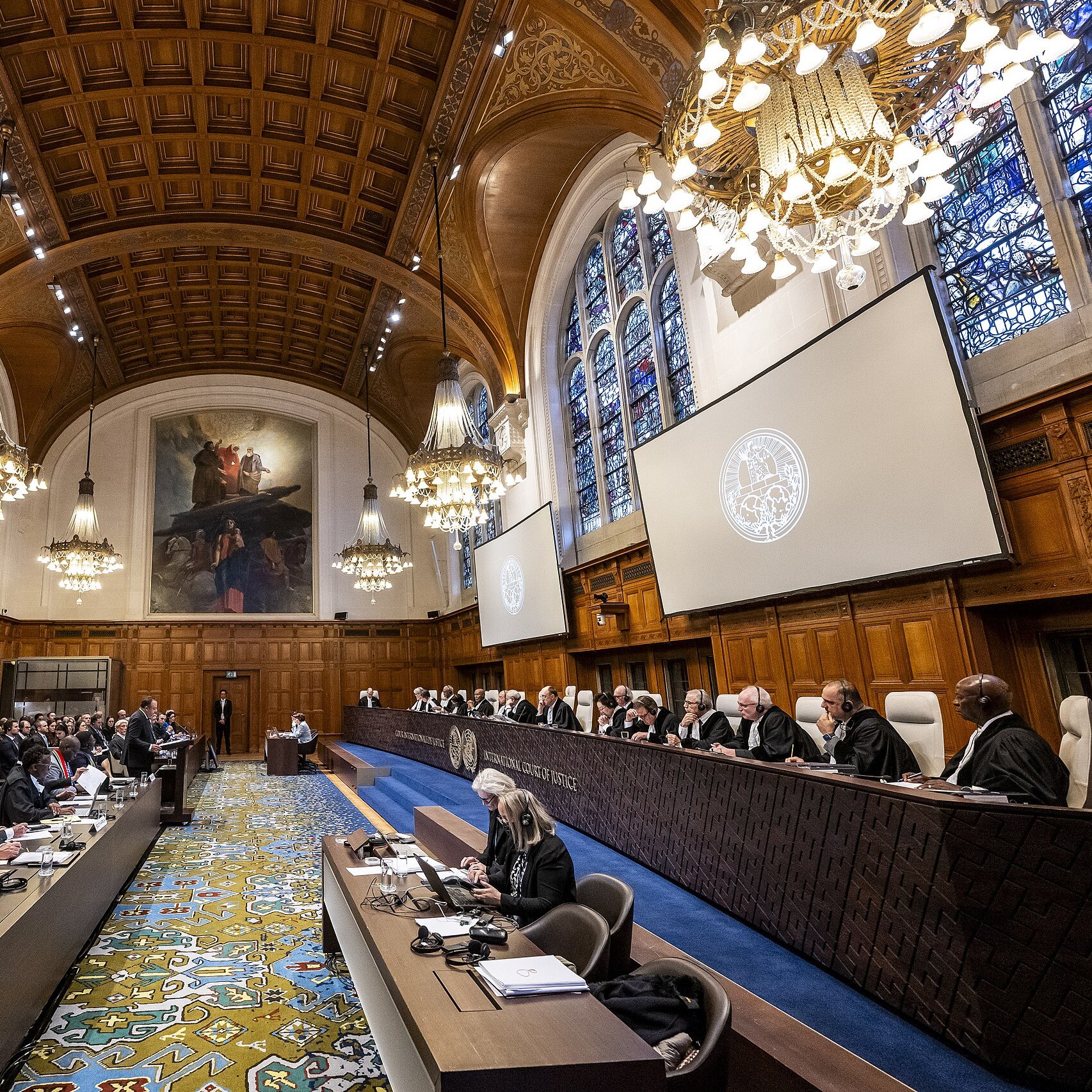 L'image montre l'intérieur d'une salle de tribunal, probablement une cour internationale. On peut y voir de nombreuses personnes assises, certaines en robes judiciaires. Au fond, des juges sont assis sur un banc élevé, entourés de décorations élégantes et de grandes fenêtres ornées de vitraux. La salle est richement décorée, avec des plafonds en bois et des lustres suspendus, créant une atmosphère solennelle et formelle. L'ensemble évoque un cadre judiciaire prestigieux.