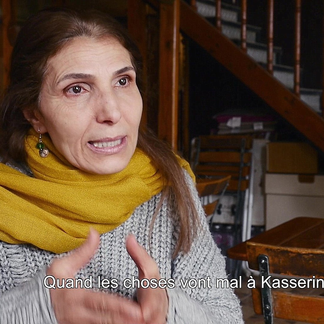 L'image montre une femme en train de parler, assise dans un environnement intérieur. Elle porte un vêtement gris avec un foulard jaune autour du cou. L'arrière-plan présente des escaliers en bois et des tables en bois, ce qui donne une ambiance chaleureuse et intime. Elle semble s'exprimer avec passion, évoquant probablement des enjeux importants liés à la ville de Kasserine.