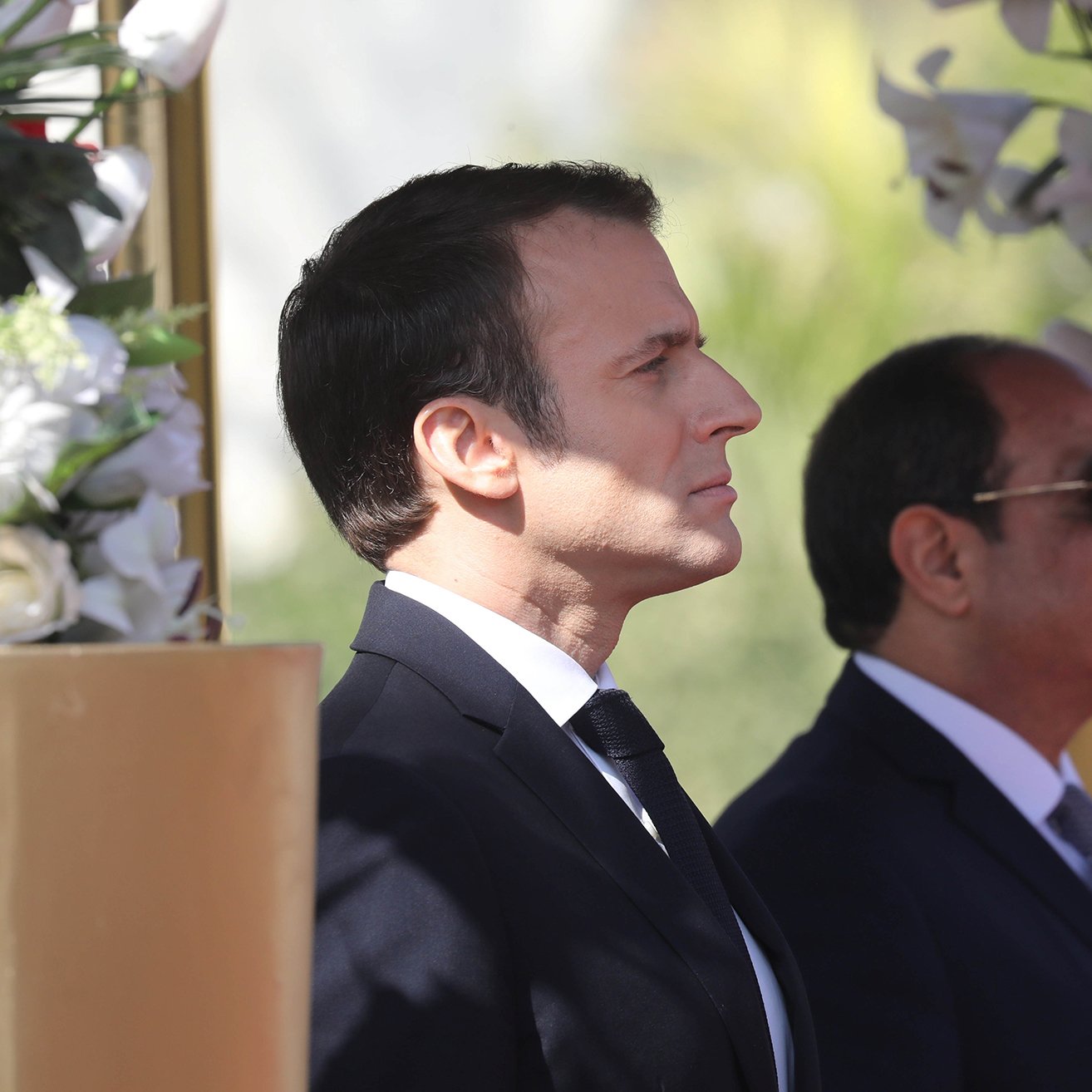 این تصویر دو مرد را در کنار هم نشان می‌دهد. شخصی در سمت چپ با موهای تیره و لباس رسمی مشکی، در حالت تفکر و جدی ایستاده است. در سمت راست، مرد دیگری با عینک آفتابی و لباس رسمی به رنگ تیره، در کنار او قرار دارد. پس‌زمینه شامل گل‌های سفید و عناصر طبیعی است که به تصویر زیبایی و شکیل بودن می‌بخشد.