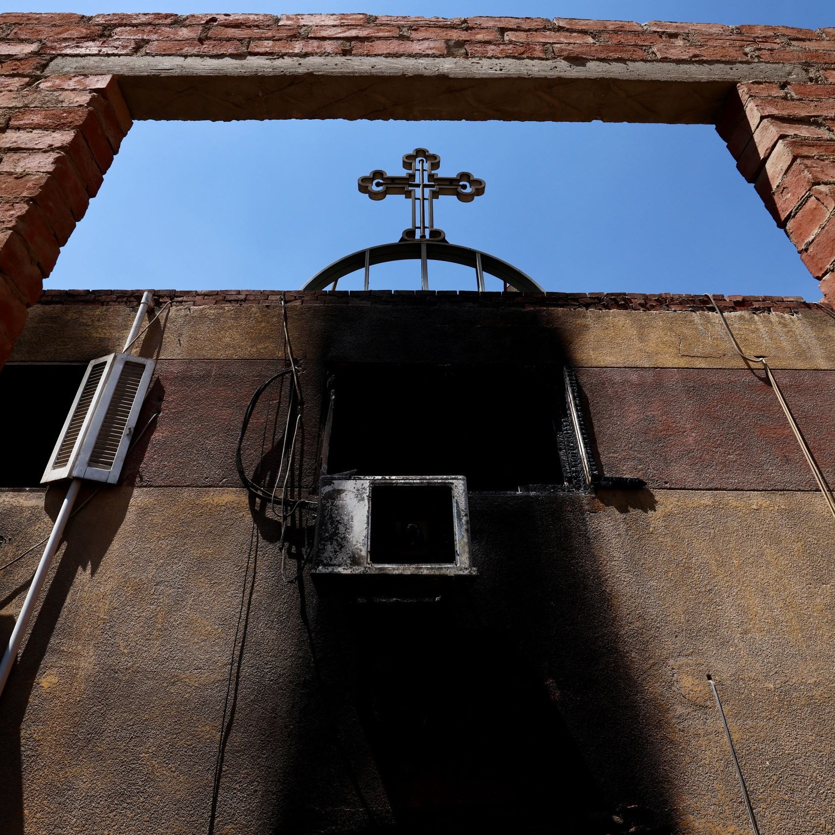 L'image montre un cadre en briques laissant entrevoir un ciel bleu, avec une croix noire au sommet d'une structure. On peut également voir des éléments d'électricité sur le mur, et une partie du mur semble avoir subi des dommages dus à un incendie, avec des marques noires. L'ensemble dégage une atmosphère de contraste entre la lumière et l'ombre, ainsi qu'une impression de désolation.