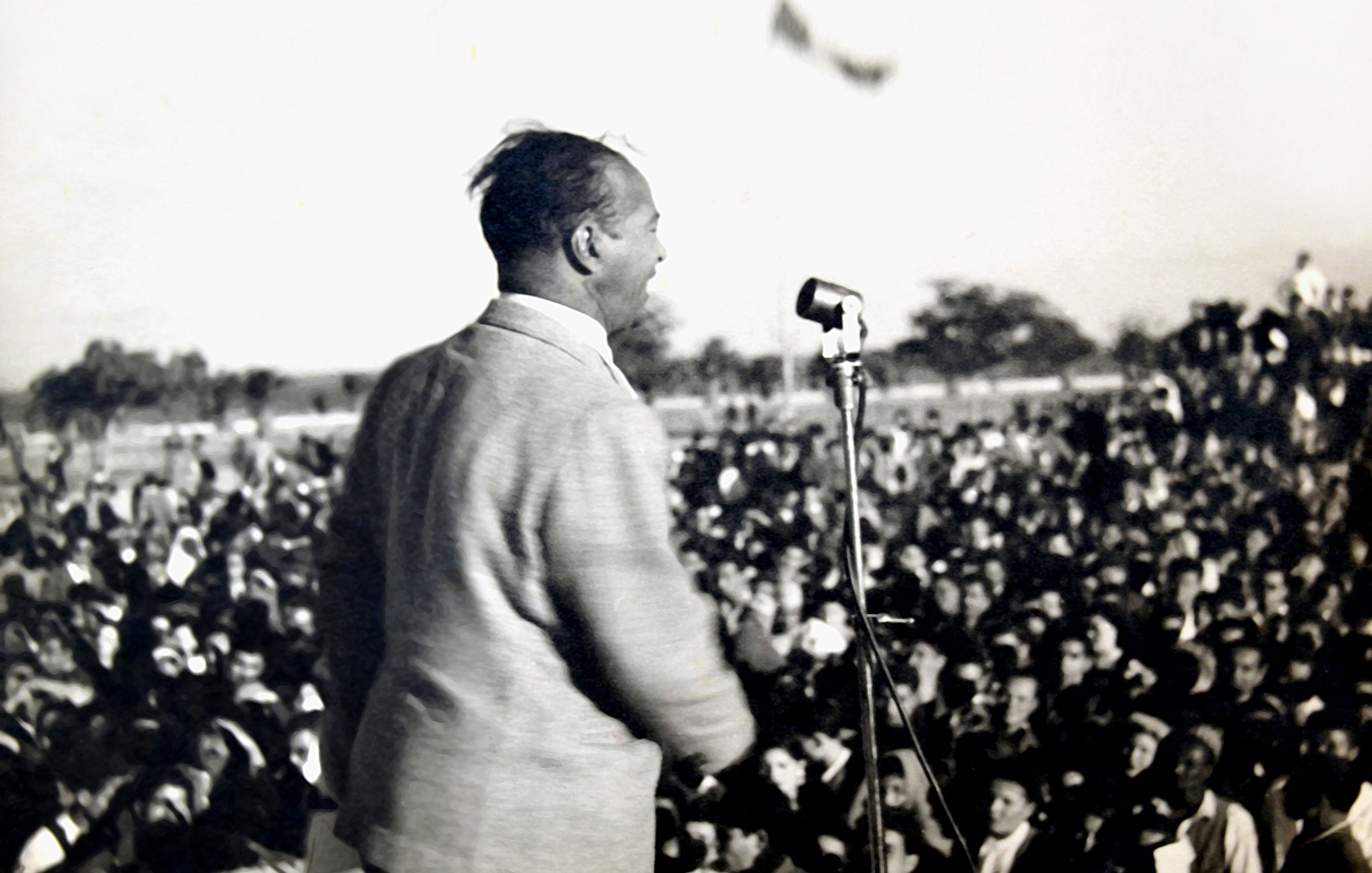 L'image montre un homme en train de parler sur une scène, devant un grand public. Il est vu de dos, et on peut voir une foule nombreuse devant lui, captivée par son discours. On distingue également un microphone sur le podium. L'atmosphère semble dynamique et engagée, probablement lors d'un événement politique ou d'un rassemblement. En arrière-plan, il y a des drapeaux, ce qui pourrait indiquer un contexte symbolique ou patriotique.