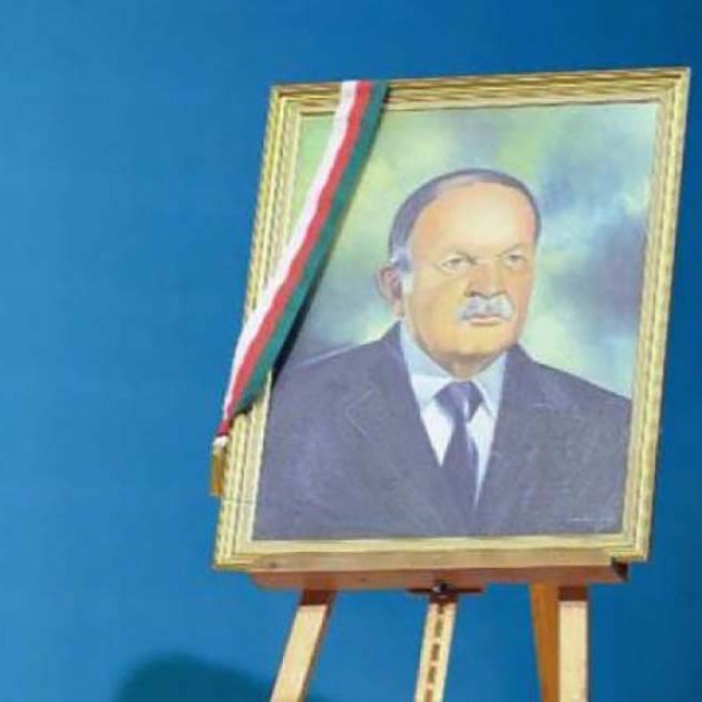 L'image montre un portrait encadré d'un homme, affiché sur un chevalet. Le fond est d'un bleu uniforme, créant un contraste avec le cadre doré du portrait. L'homme dans l'image a une expression sérieuse et porte un costume, accompagné d'un insigne ou d'un drapeau sur le cadre, possiblement représentant un symbole national ou officiel.