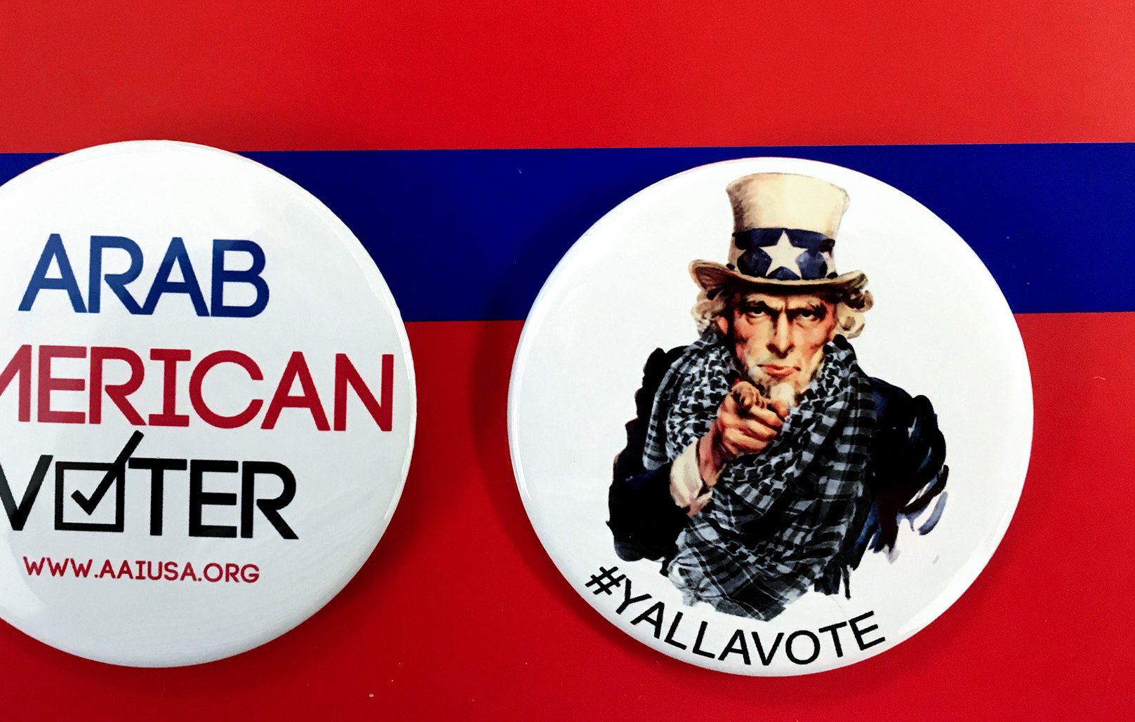 L'image montre deux badges sur un fond rouge avec une bande bleue. Le premier badge mentionne "ARAB AMERICAN VOTER" avec un symbole de coche, tandis que le deuxième badge représente un personnage qui ressemble à l'oncle Sam, avec le texte "#YALLAVOTE". Ces badges mettent en avant l'importance du vote et de l'engagement civique, en particulier au sein de la communauté arabe-américaine.