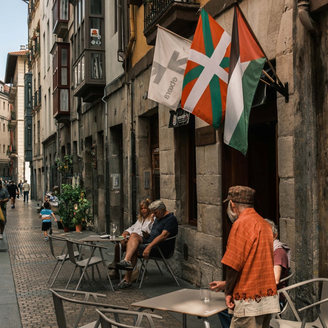 Vicolo affollato con tavolini all'aperto e bandiere che sventolano.