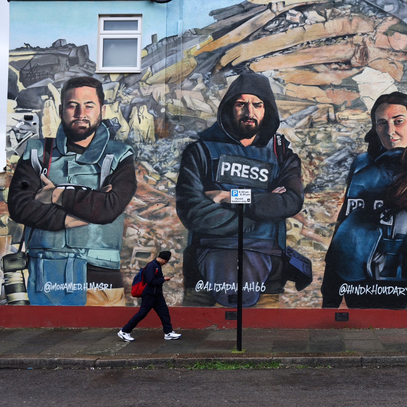 این تصویر یک دیوارنگاری بزرگ را نشان می‌دهد که چهار نفر را به تصویر کشیده است. این افراد در لباس‌های مخصوص خبرنگاری قرار دارند و با ظاهری جدی و ثابت ایستاده‌اند. در پس‌زمینه، ویرانه‌هایی دیده می‌شود که نشان‌دهنده‌ی یک حادثه یا بحران است. یکی از افرادی که در تصویر قرار دارد، لباس مخصوص "پرس" (خبرنگار) به تن دارد و همگی به دوربین نگاه می‌کنند. در جلوی دیوارنگاری، فردی در حال عبور است. این تصویر به نظر می‌رسد که به اهمیت کار خبرنگاران در پوشش اخبار بحران‌ها اشاره دارد.