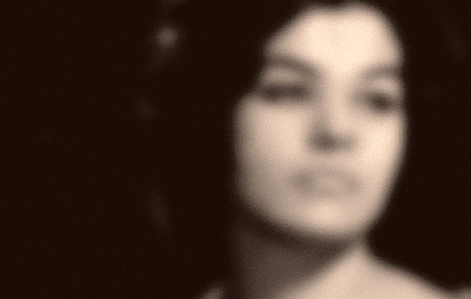 L'image montre une femme dont le visage est flou. Elle a les cheveux longs et bouclés, et semble perdre un regard pensif ou distant. L'arrière-plan est sombre, ce qui ajoute à l'atmosphère de mystère et de nostalgie de la photo. Son expression laisse entrevoir des émotions profondes, mais les détails précis restent indéfinis.