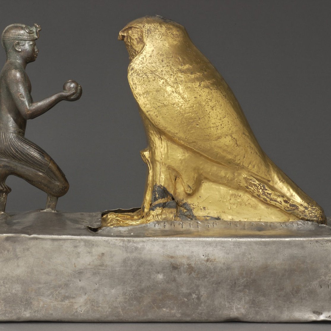 L'image représente une sculpture en métal, où l'on peut voir un personnage humain à genoux tenant un objet dans ses mains, face à un grand hibou doré. Le personnage est sculpté de manière détaillée, avec des traits qui évoquent un style ancien, probablement lié à l'Égypte ou à une culture similaire. Le hibou, de couleur dorée, symbolise peut-être la sagesse ou un aspect divin. La base de la sculpture est en métal argenté, offrant un contraste intéressant avec la brillance du hibou doré. Cette œuvre semble revêtir une signification symbolique ou religieuse.