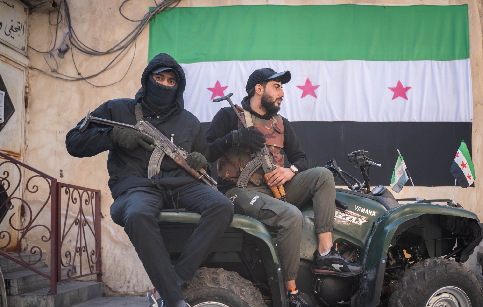 Deux hommes assis sur un quad, l'un tenant une arme, l'autre un drapeau. En arrière-plan, un mur avec un grand drapeau portant des étoiles rouges sur fond vert et noir. L'ambiance semble militaire et tendue.