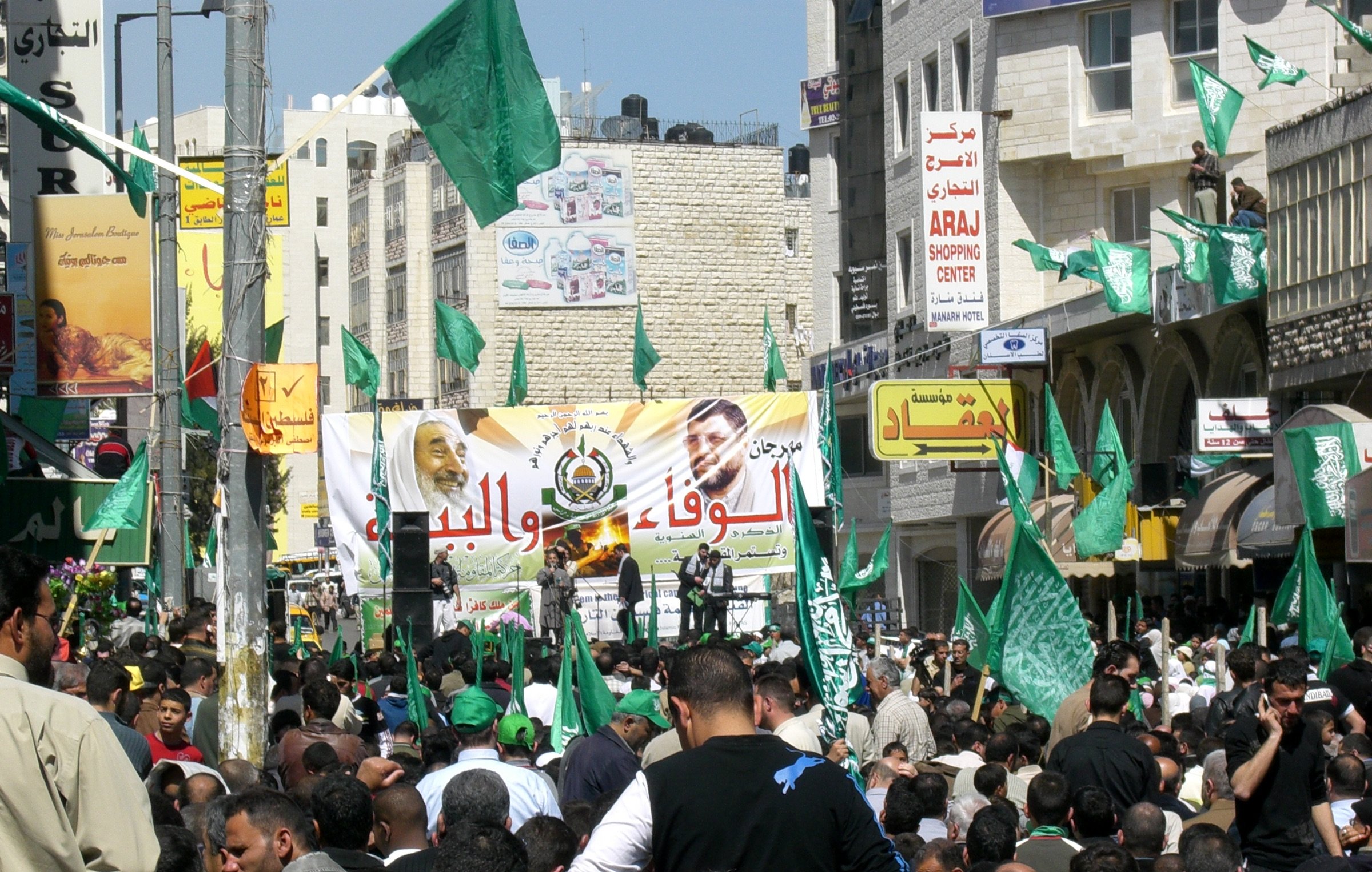 L'image montre une foule dense rassemblée dans une rue, manifestant avec des drapeaux verts. Au fond, il y a une grande banderole, probablement portant des messages politiques. Les gens semblent enthousiastes et engagés, et l'environnement est animé avec des bâtiments et des enseignes visibles autour. Les drapeaux verts peuvent indiquer l'association avec un mouvement ou un parti spécifique.