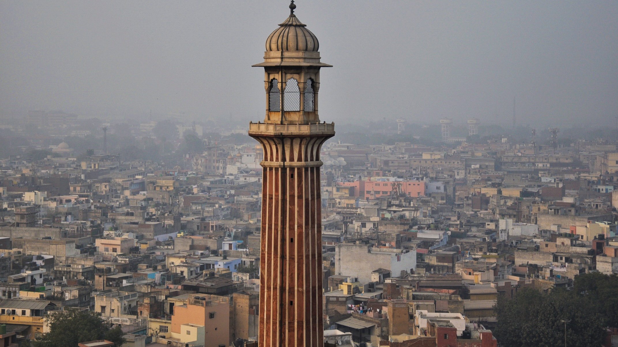 L'image montre un paysage urbain avec une tour élancée qui se dresse au milieu de nombreux bâtiments. La tour a une architecture distinctive, avec des motifs ornés et un dôme en haut. En arrière-plan, on peut voir une vaste étendue de maisons et de structures, témoignant d'une villeDense. L'atmosphère semble légèrement brumeuse, suggérant une pollution ou une humidité ambiante. Les couleurs des bâtiments varient, ajoutant une touche de diversité à l'ensemble de la scène.