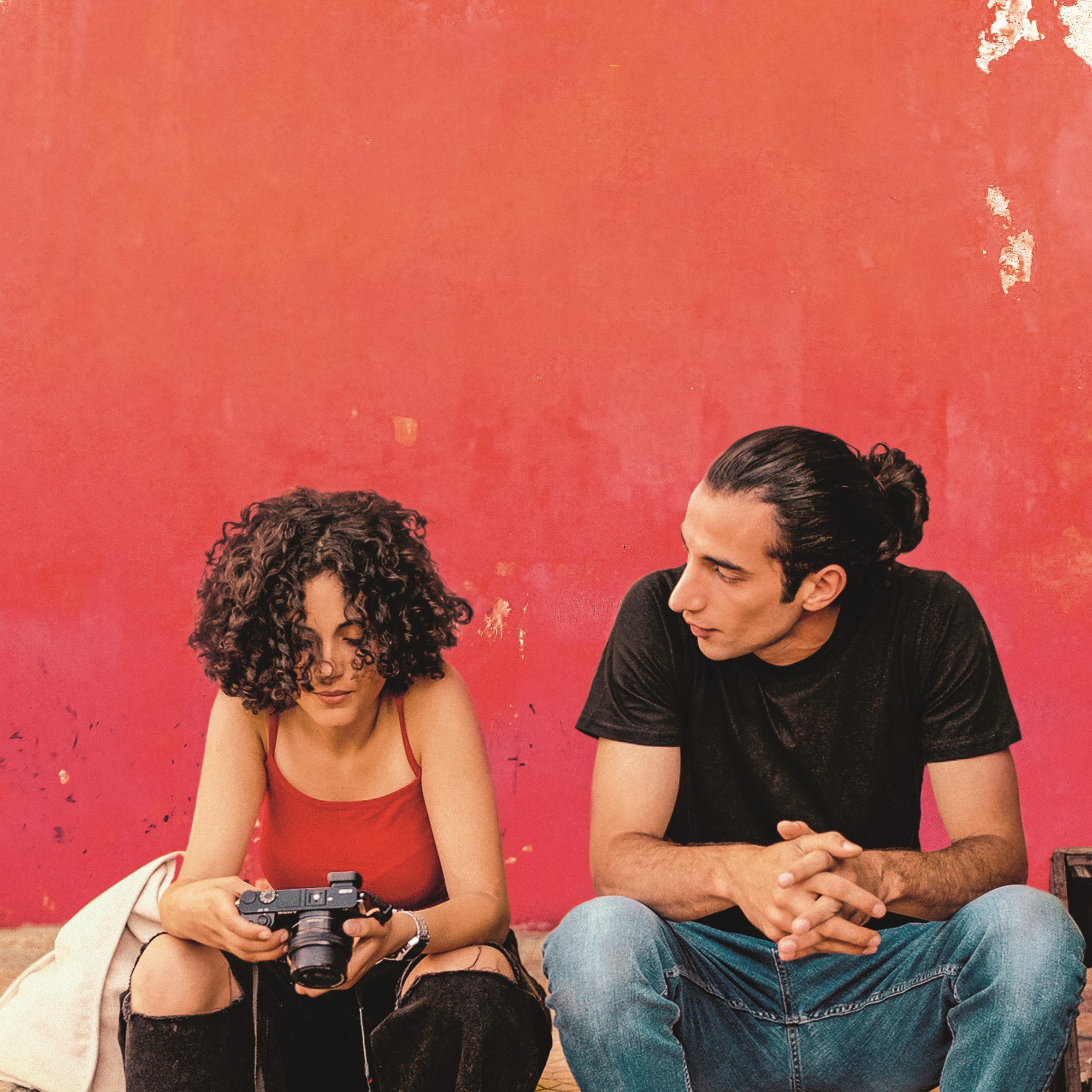 L'image montre deux personnes assises devant un mur rouge écaillé. La première personne, une femme aux cheveux bouclés, tient un appareil photo et semble concentrée sur son écran. Elle porte un débardeur rouge et un gilet clair. À côté d'elle, un homme avec des cheveux longs attachés en chignon la regarde attentivement. Il est vêtu d'un t-shirt noir et de jeans. L'atmosphère paraît détendue et intime, comme s'ils étaient en pleine conversation ou en train de partager un moment.
