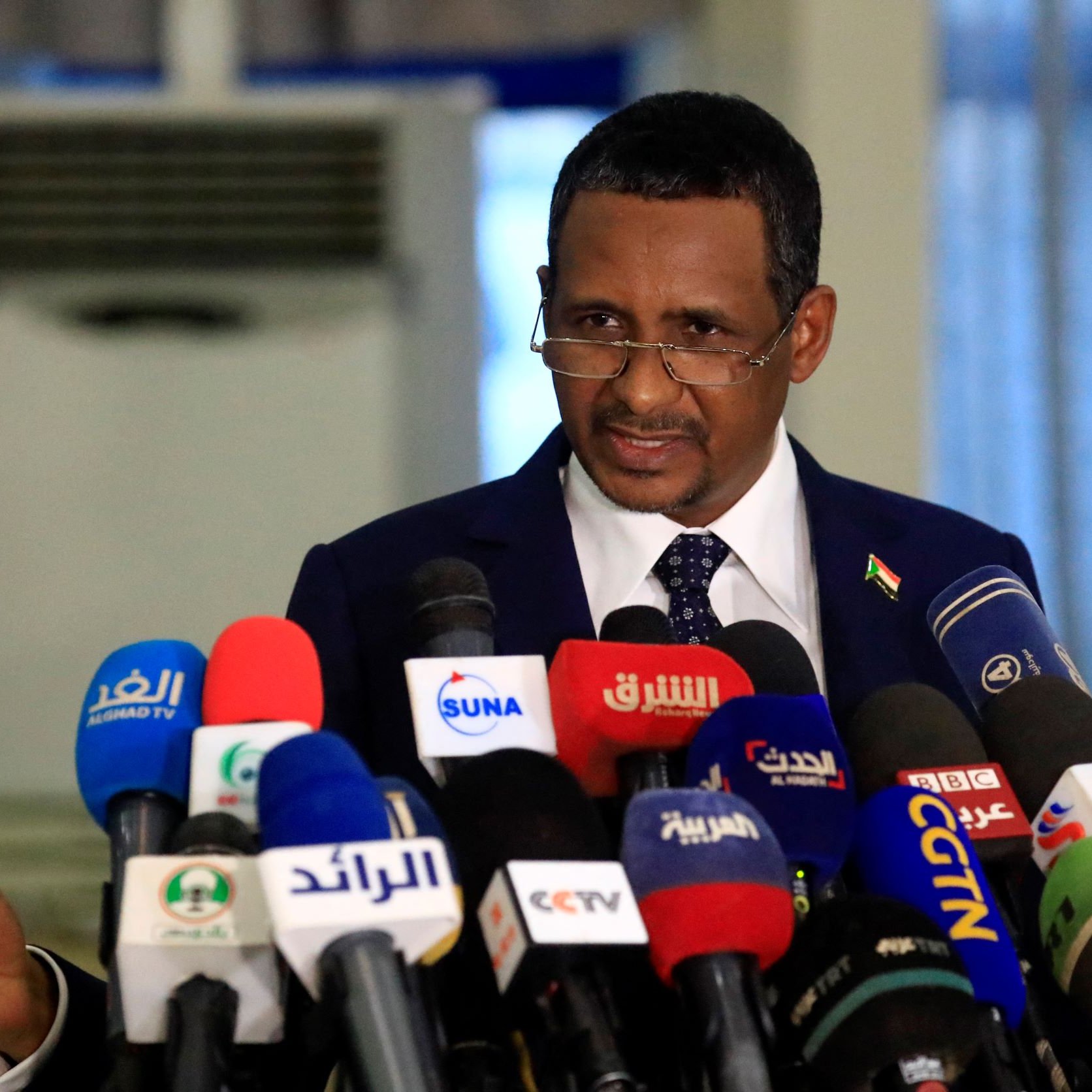 در این تصویر، مردی در حال صحبت کردن در جلوی میکروفن‌های زیادی است. او لباس رسمی به تن دارد و به نظر می‌رسد در حال ارائه یک سخنرانی یا بیانیه است. در پس‌زمینه، تعدادی دوربین و میکروفون وجود دارد که نشان‌دهنده جوی رسانه‌ای و توجه عمومی به صحبت‌های اوست. فضا به‌نظر رسمی و جدی می‌رسد.