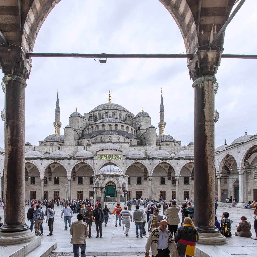 این تصویر نمایی از مسجد سلطان احمد (مسجد آبی) در استانبول را نشان می‌دهد. در foreground (پیش‌زمینه)، حیاط بزرگ مسجد با ستون‌های بلند و قوس‌های زیبای آن دیده می‌شود. مردم در حال تردد و بازدید از این مکان تاریخی هستند. در پس‌زمینه، ساختمان اصلی مسجد با گنبد بزرگ و مناره‌های بلندش خودنمایی می‌کند. آسمان نسبتاً ابری است و اجسام و ساختمان‌ها در نور ملایم قرار دارند. جو کلی تصویر شلوغ و زیبا است که تاریخ و فرهنگ غنی این مکان را منتقل می‌کند.