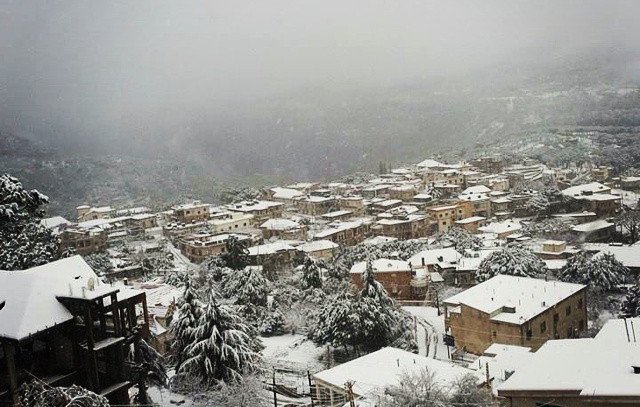 L'image montre un paysage hivernal où une petite ville est recouverte de neige. Les toits des maisons, en grande partie en bois, sont également blancs, créant une atmosphère paisible et froide. On peut apercevoir des arbres chargés de neige et des collines en arrière-plan, le tout enveloppé par une brume légère, ajoutant à la sérénité de la scène. La vue donne une impression de calme et d'isolement, typique des régions montagneuses en hiver.