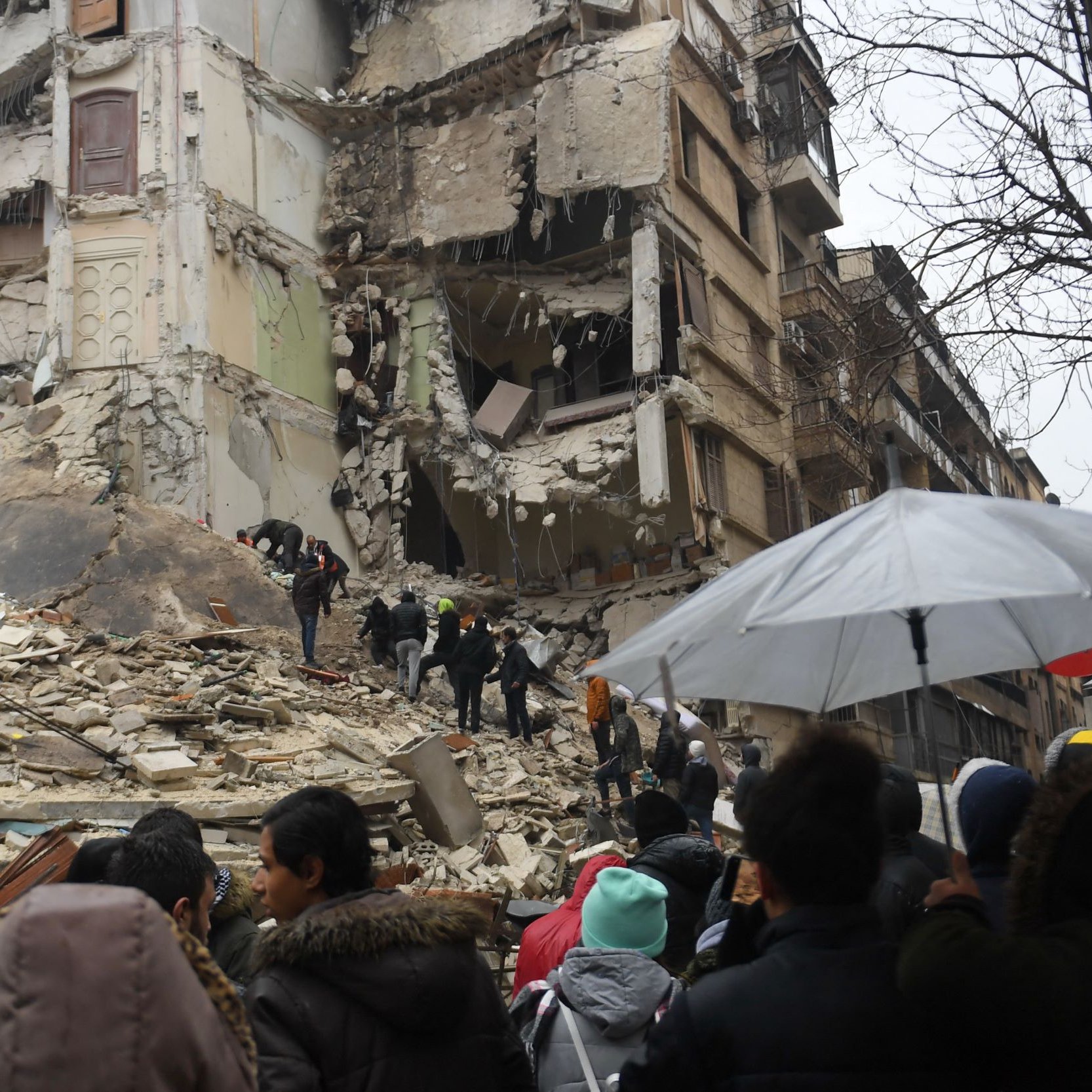 L'image montre une scène de destruction après un tremblement de terre. On peut voir un bâtiment gravement endommagé, avec des murs effondrés et des débris éparpillés. Au premier plan, une foule de personnes semble se rassembler, regardant les secours qui travaillent. Certains porteurs d'ombrelles se protègent de la pluie. L'atmosphère est sinistre, et l'on ressent une forte émotion face à la destruction causée par la catastrophe.