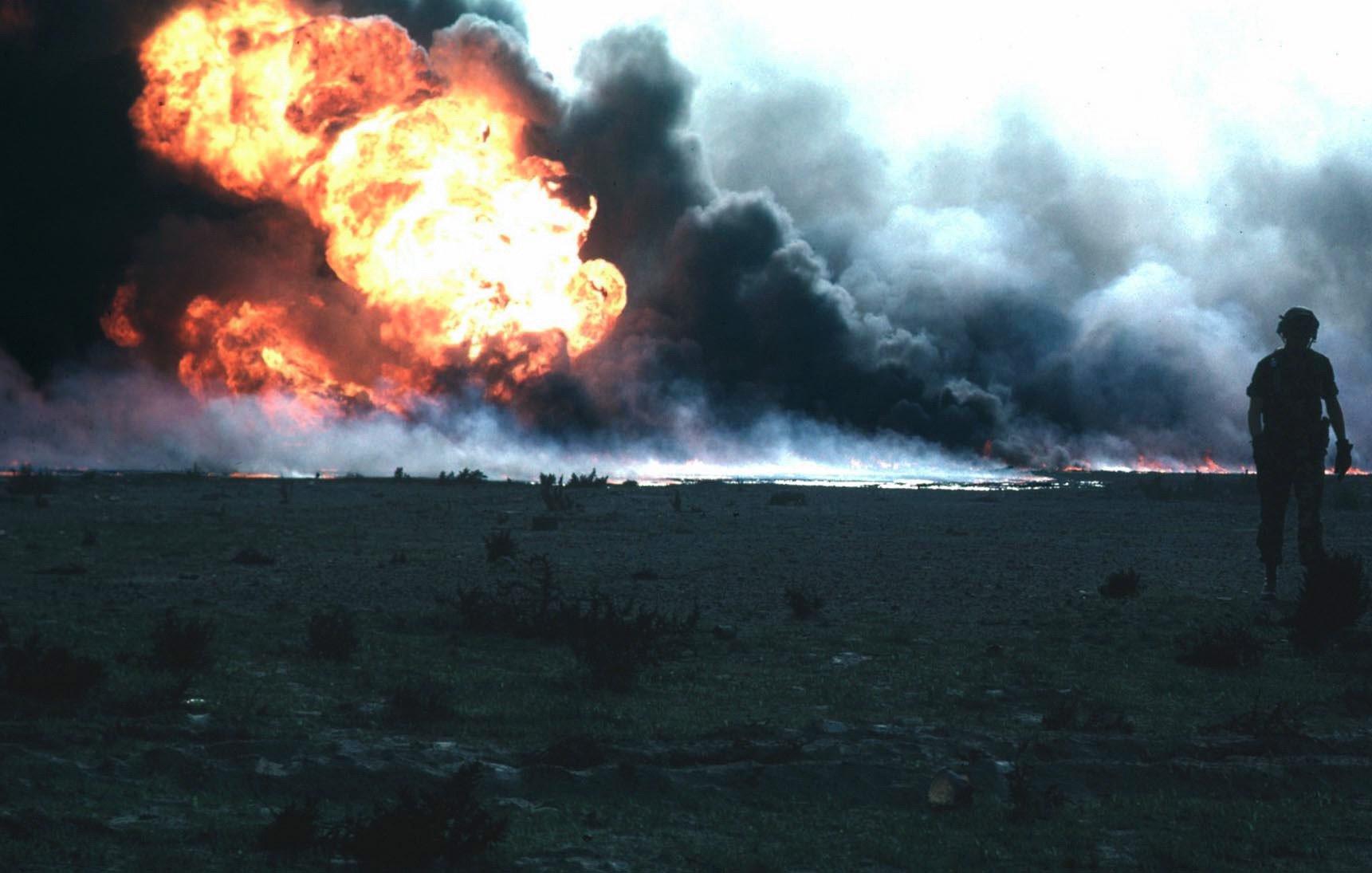 L'image montre une scène de destruction avec un grand incendie au loin. Des flammes vives jaillissent dans un nuage de fumée noirâtre. Au premier plan, deux silhouettes de soldats sont visibles, marchant côte à côte. Le paysage semble aride, avec peu de végétation. L'atmosphère est sombre et dramatique, soulignant l'impact de l'explosion.