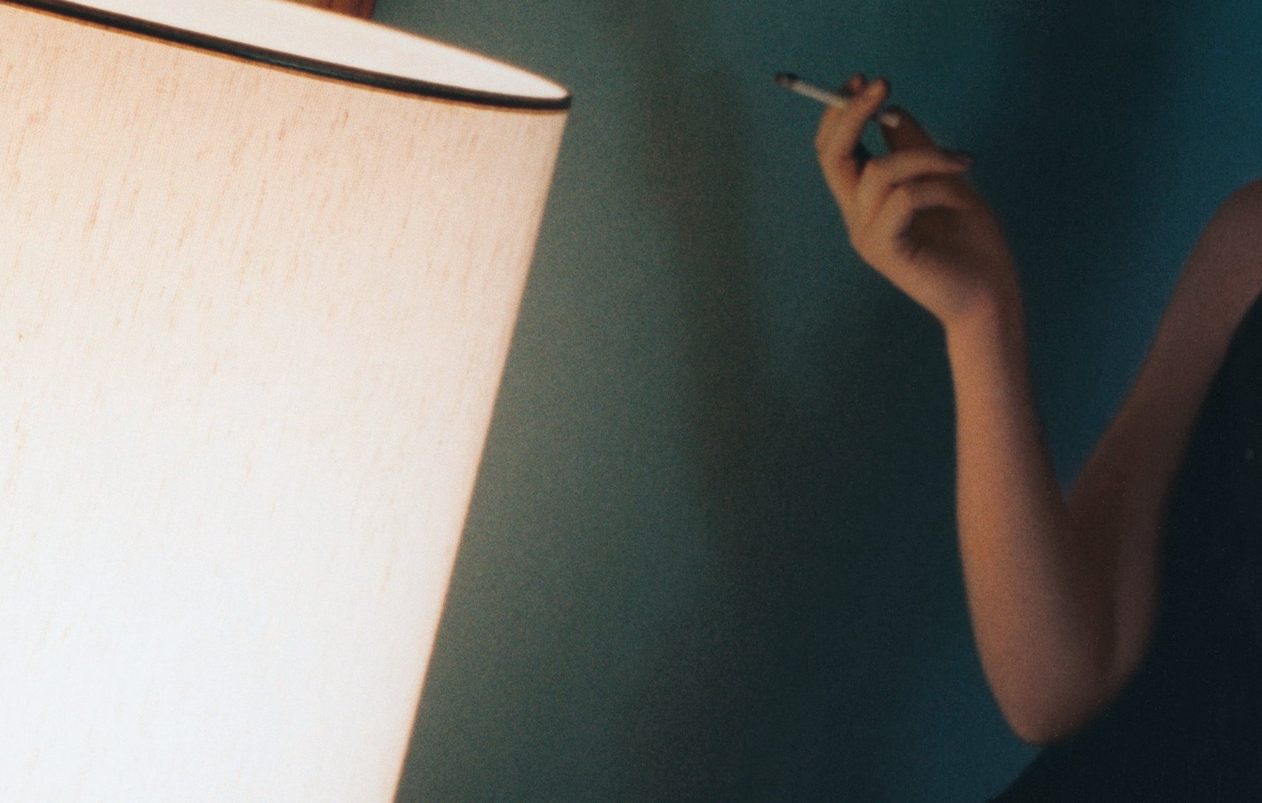 L'image montre un intérieur avec une lumière douce émise par une lampe. Une personne, située à gauche, tient un objet à la main qui semble être une cigarette. La décoration murale est d'une teinte douce, créant une atmosphère intime et relaxante. La lumière de la lampe joue sur les surfaces, ajoutant une ambiance chaleureuse à la scène.