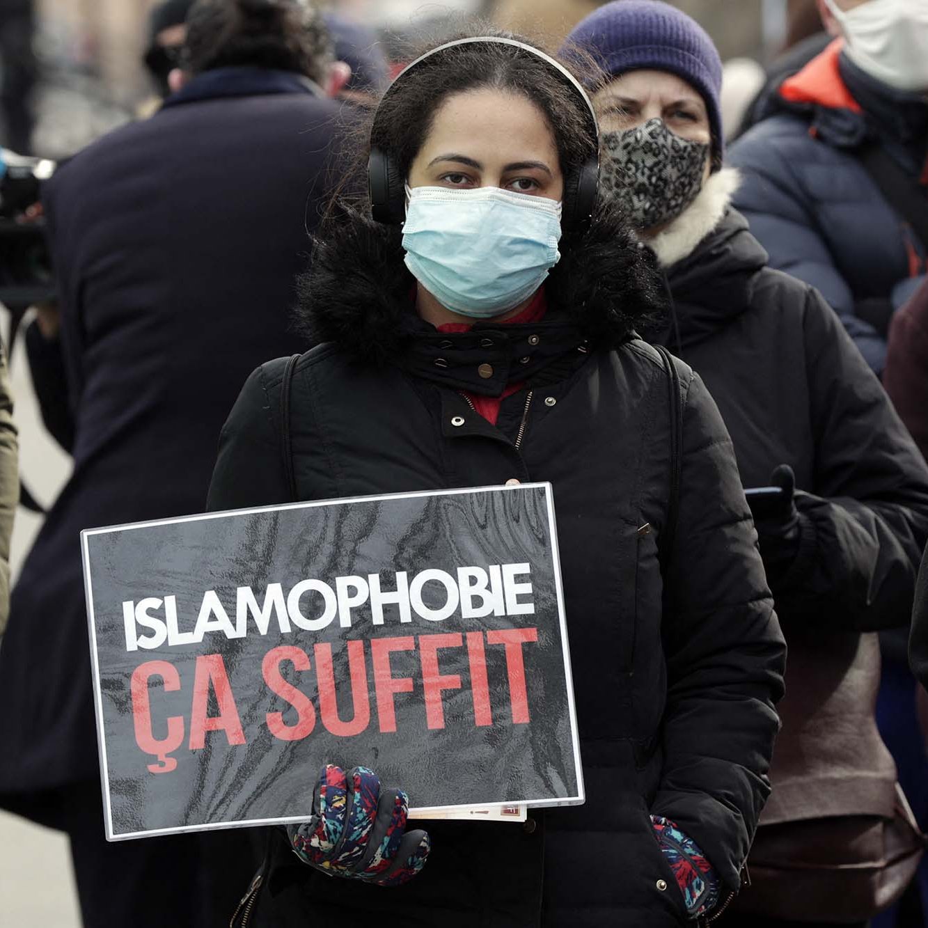 L'image montre une femme portant un masque, se tenant au milieu d'une foule. Elle tient une pancarte sur laquelle est écrit "ISLAMOPHOBIE ÇA SUFFIT". Les personnes autour d'elle semblent également s'engager dans une manifestation, témoignant d'un soutien à la cause qu'elle défend. L'atmosphère est sérieuse et engagée, reflétant des préoccupations sociales.