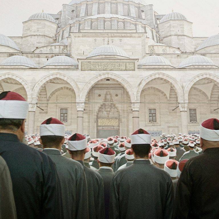 L'image montre un grand groupe de personnes, probablement des religieux ou des fidèles, qui se tiennent debout, tournés vers un imposant bâtiment architectural qui semble être une mosquée. Les hommes portent des vêtements similaires, souvent dans des tons sombres, et certains portent des turbans ou des chapeaux à pompons rouges. L'ambiance semble solennelle et respectueuse, suggérant un moment de prière ou de rassemblement communautaire. Les détails architecturaux de la mosquée, avec ses coupoles et arches majestueuses, ajoutent à l'imposante scène.