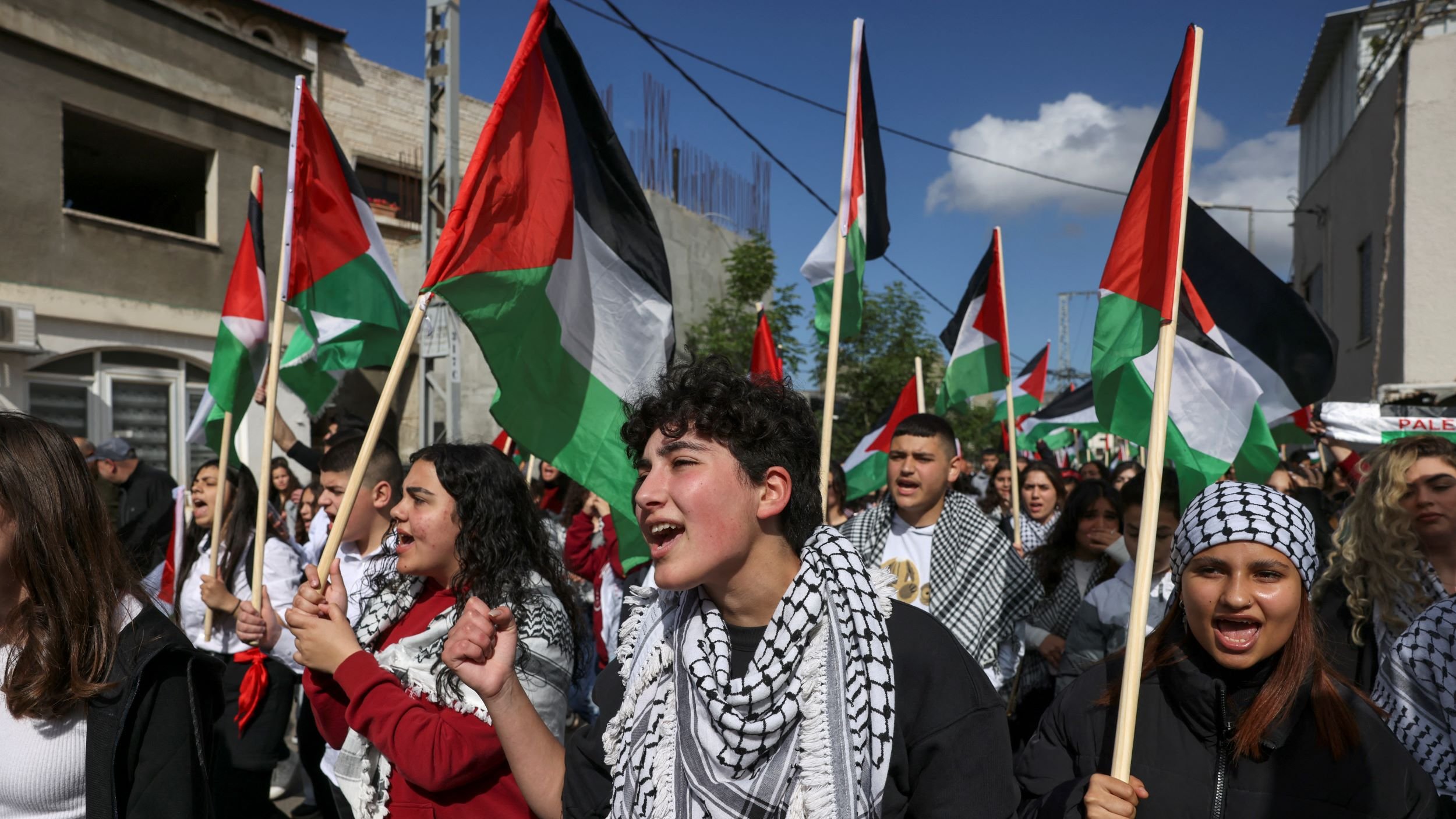 L'immagine mostra un gruppo di persone che partecipano a una manifestazione. Molti manifestanti tengono in mano bandiere palestinesi, caratterizzate da strisce nere, bianche e verdi e da un triangolo rosso. Alcuni di loro indossano kefiah, tradizionali sciarpe palestinesi. Le espressioni sui loro volti trasmettono passione e determinazione mentre cantano o gridano slogan, contribuendo a creare un'atmosfera di unità e attivismo. Sullo sfondo, si intravedono edifici e il cielo è parzialmente nuvoloso.