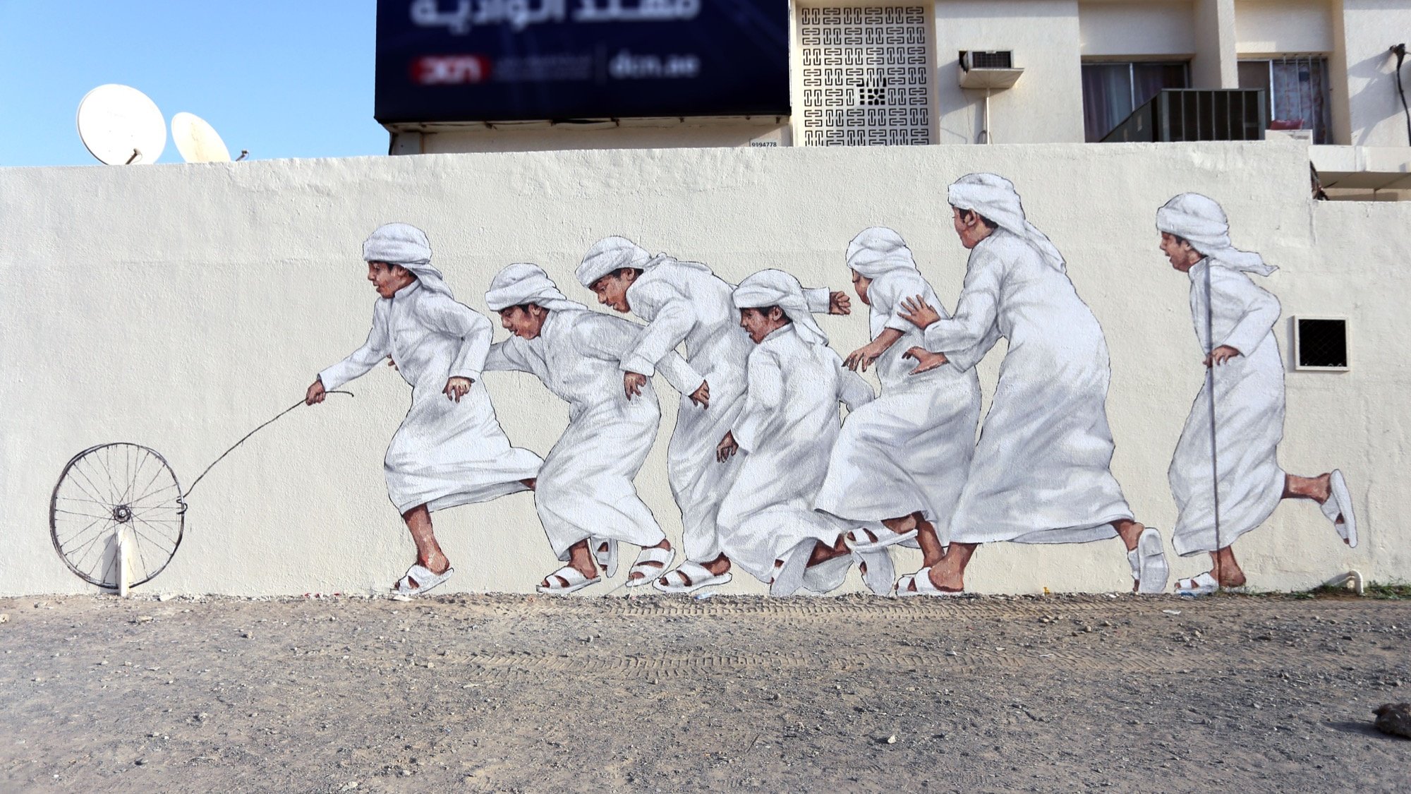 L'image montre une fresque murale représentant plusieurs enfants en vêtements traditionnels blancs. Ils semblent courir joyeusement, certaines figures tenant un fil ou une roue, ce qui évoque un jeu. Le décor est d'un style urbain, avec un fond minimaliste et une ambiance dynamique. L'art mural met en valeur la culture et la joie de l'enfance.