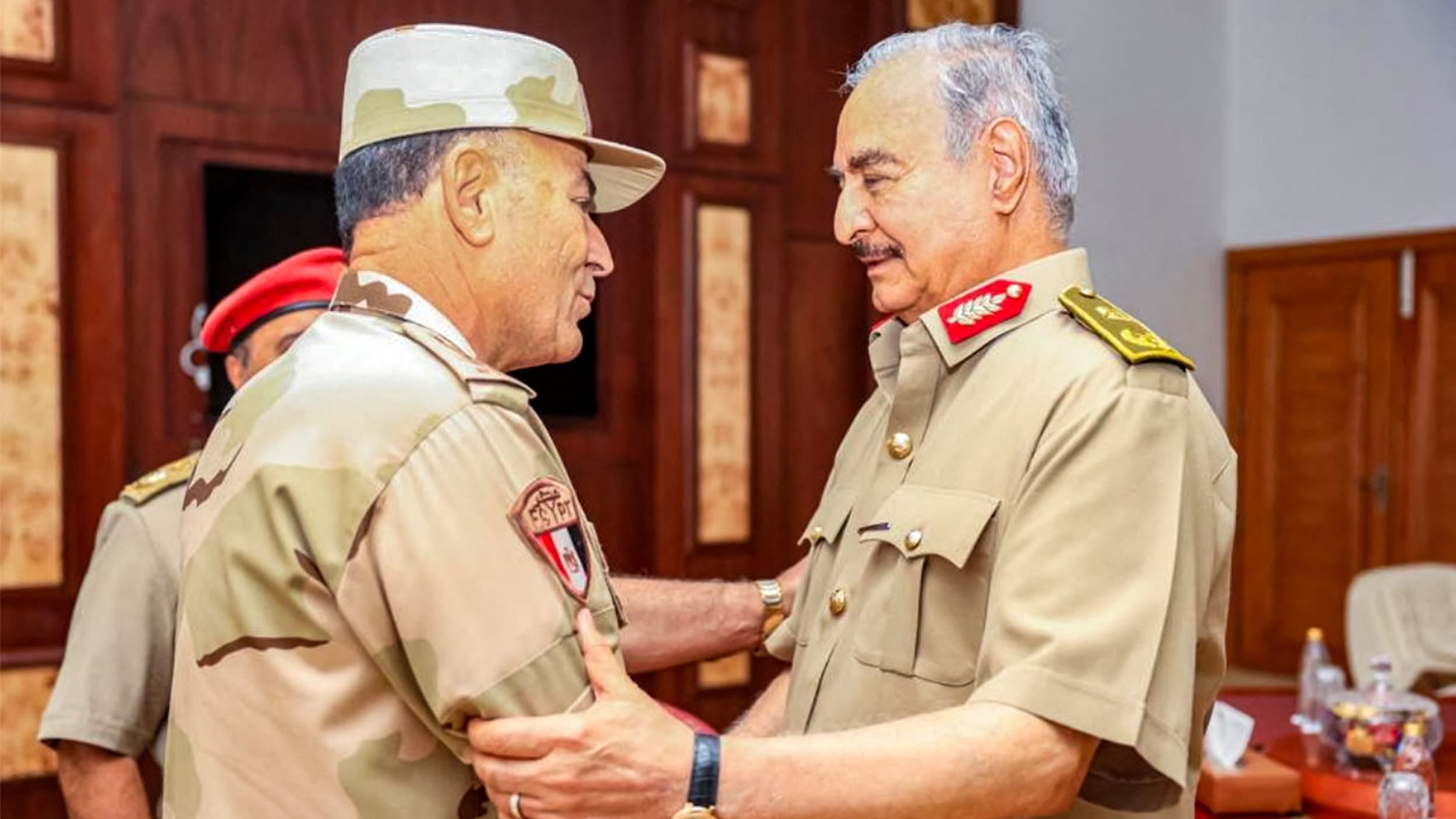 L'image montre deux militaires en uniforme, se tenant face à face et en train de discuter. L'un d'eux porte un chapeau militaire et un uniforme beige avec des motifs camouflage, tandis que l'autre est habillé en uniforme militaire beige et porte des décorations sur son épaule. Ils semblent avoir une conversation amicale et posent dans un environnement intérieur, avec des tables et des chaises visibles en arrière-plan. L'atmosphère semble informelle, suggérant une réunion ou un échange entre officiers.