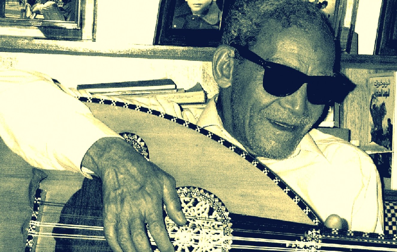 L'image montre un homme âgé portant des lunettes de soleil, assis et jouant de l'oud, un instrument de musique traditionnel. Il semble concentré sur sa musique, avec un sourire léger. En arrière-plan, on aperçoit des photos encadrées accrochées au mur, ce qui pourrait suggérer un environnement familial ou personnel. L'éclairage donne une ambiance nostalgique à la scène.