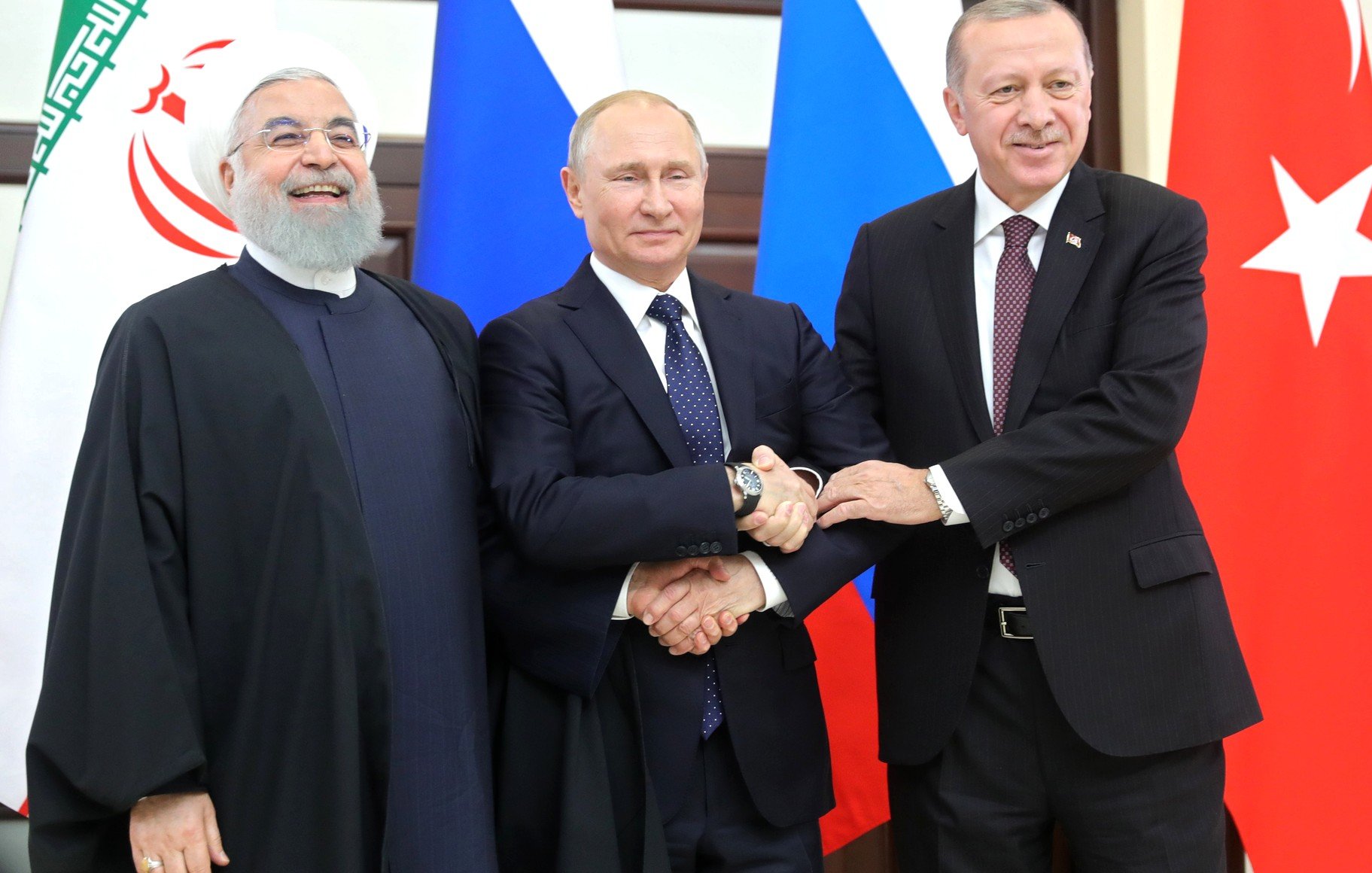 في الصورة، يظهر ثلاثة شخصيات سياسية بارزة. الشخص على اليسار يرتدي لباساً تقليدياً إسلامياً وله لحية. في الوسط، يظهر شخص يرتدي بدلة رسمية وهو يبدو كزعيم دولة. على اليمين، يظهر شخص آخر يرتدي بدلة أيضاً، وهو يبتسم. الثلاثة يقفون معاً ويبدو أنهم يتصافحون أو يضعون أذرعهم على بعضهم البعض. خلفهم توجد أعلام دول مختلفة، مما يشير إلى اجتماع رسمي أو قمة.