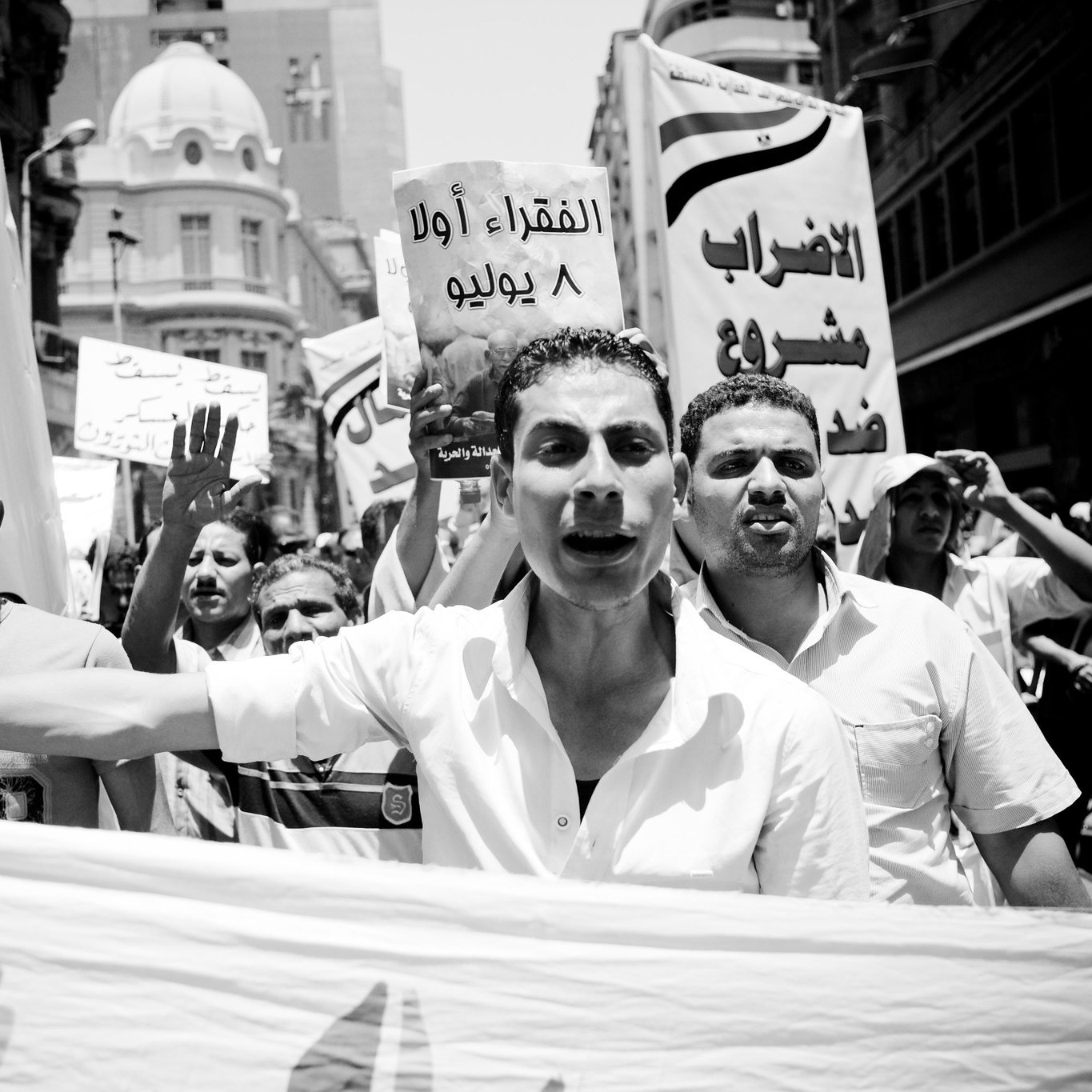 The image depicts a black and white scene of a protest or demonstration. A group of people is marching together, holding banners and signs. One person in the foreground appears to be passionately speaking or chanting, while others raise their fists or shout in the background. The signs seem to convey messages related to social or political issues, with at least one sign written in Arabic. The overall atmosphere suggests urgency and activism, capturing a moment of collective expression and dissent.