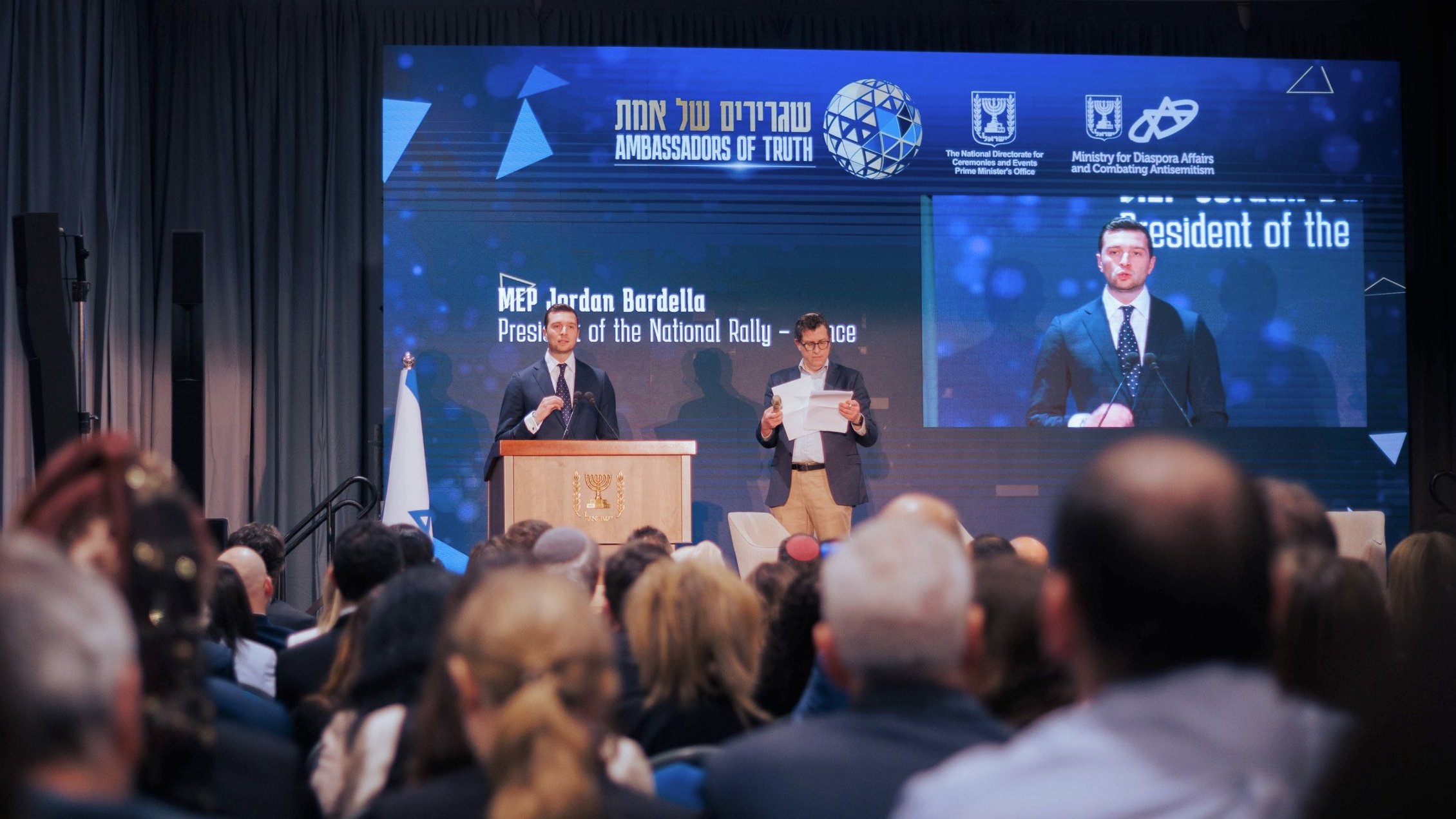 الصورة تظهر حدثًا رسميًا حيث يتحدث متحدث أمام جمهور كبير. على المنصة، يوجد شخص يحمل ورقة، ويبدو أن المتحدث يقرأ من نص. هناك خلفية تعرض شعارًا ومعلومات عن الفعالية. الأجواء تبدو جدية والر audience مهتم.