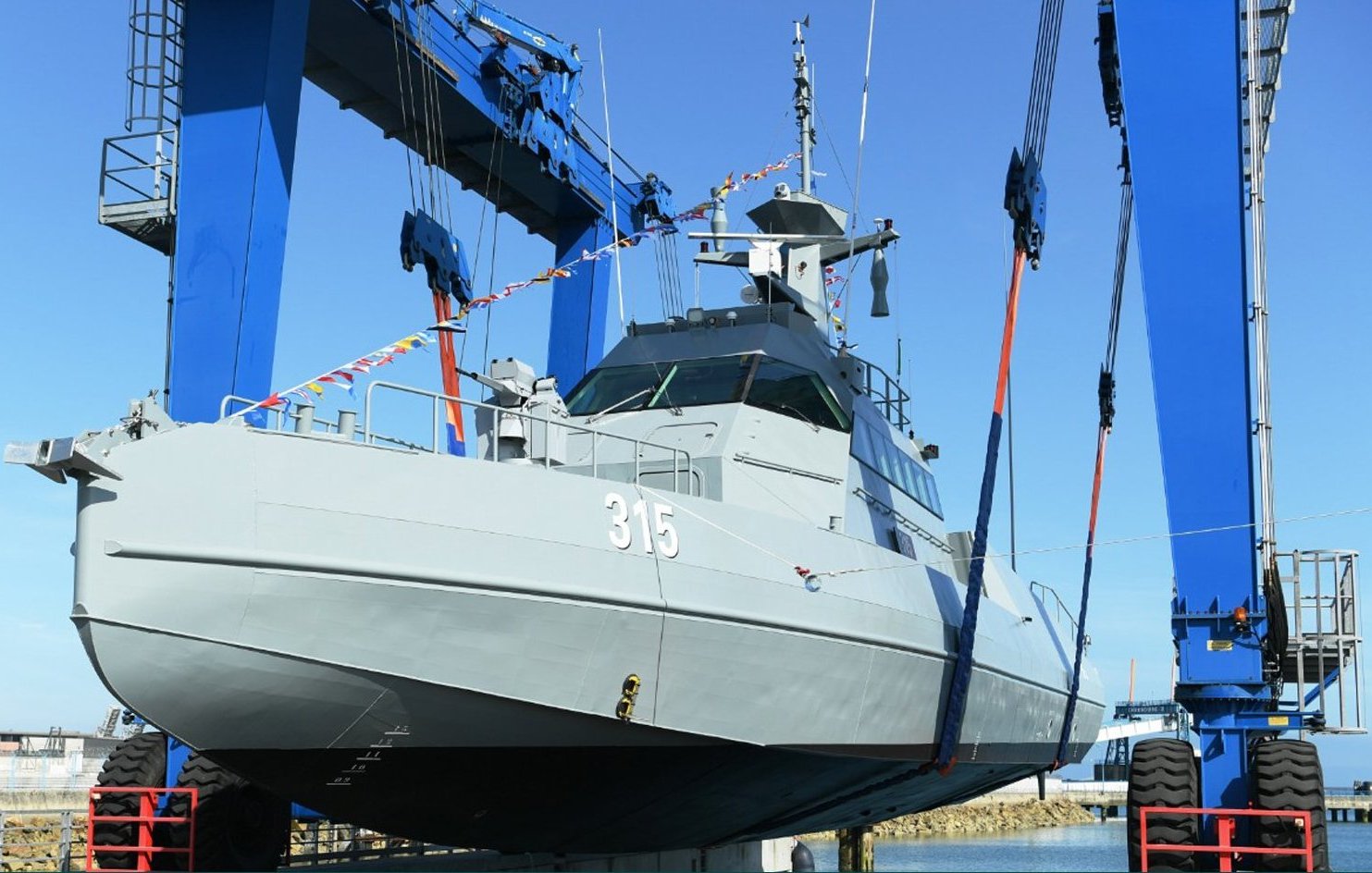 L'image montre un navire militaire en cours de manutention. Il est positionné sur un quai, soutenu par des grues bleues qui le soulèvent avec des sangles. Le bateau est de couleur grise et porte le numéro 315. Le ciel est clair, suggérant une journée ensoleillée. Cette scène évoque des opérations de maintenance ou de construction navale.