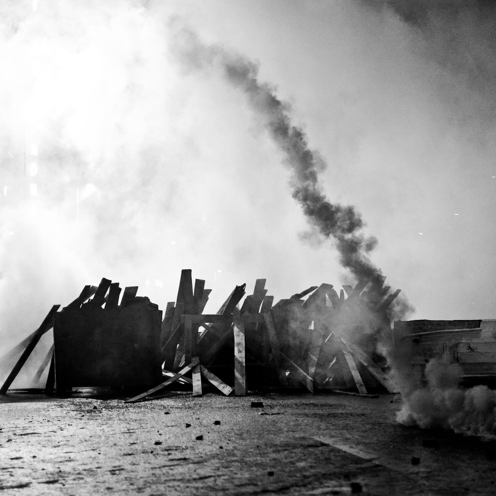 L'image montre une scène de tension, probablement associée à une manifestation ou à des troubles civils. On y voit des barricades faites de planches de bois, qui semblent être disposées sur une route. Une épaisse fumée s'élève dans l'air, créant une atmosphère sombre et chaotique. La monochromie de la photo ajoute à l'intensité de la scène, soulignant le contraste entre les ombres et la lumière. Des éléments comme des fumigènes ou des projectiles peuvent également être présents, suggérant une confrontation entre manifestants et forces de l'ordre. L'ensemble évoque une situation de conflit et de désobéissance civile.