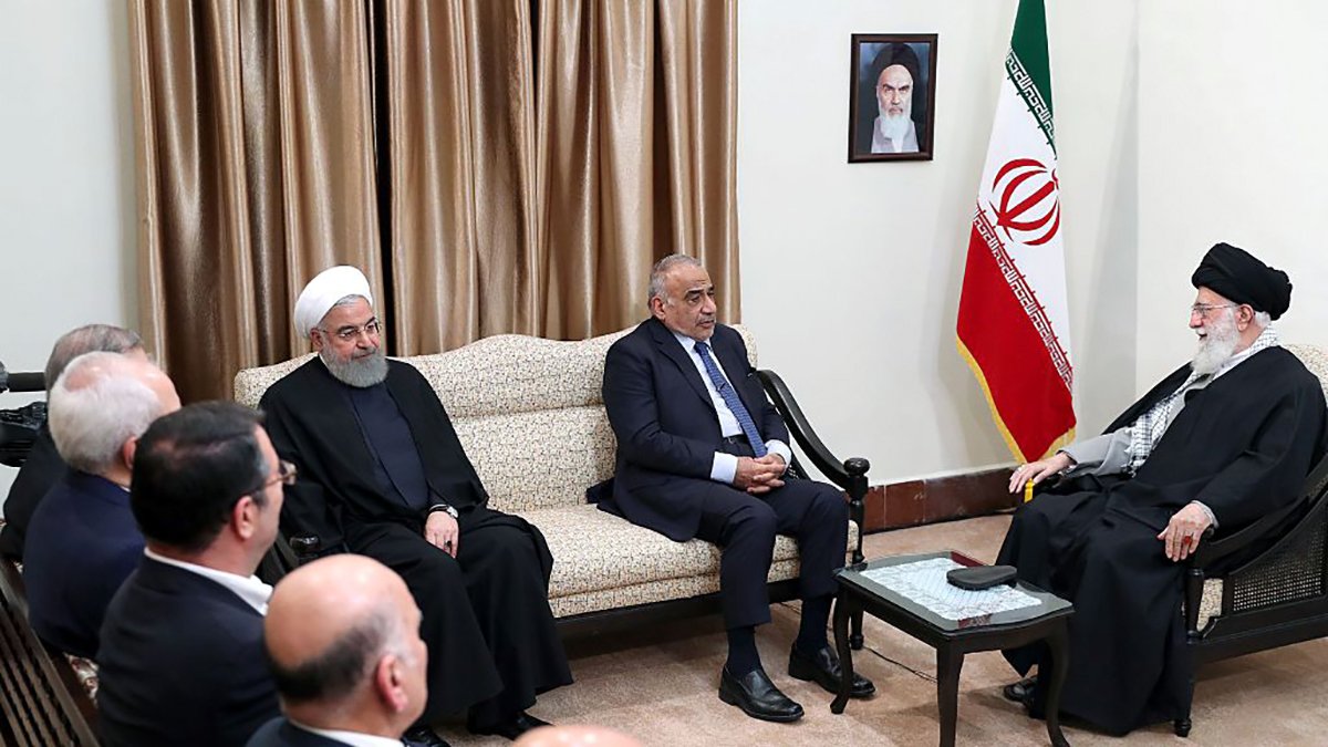 در این تصویر، چند نفر در یک اتاق رسمی نشستهاند. شخصی در سمت راست تصویر با لباس مشکی و عمامه، رهبر جمهوری اسلامی ایران است. در مجاورت او، فرد دیگری نشسته که به نظر میرسد مرتبط با مقامات دولتی باشد. در سمت چپ، افراد دیگری در حال گوش دادن به صحبتها هستند. دیوارها با پردههای قهوهای تزیین شده و پرچم جمهوری اسلامی ایران در پسزمینه دیده میشود. یک تصویر از بنیانگذار جمهوری اسلامی نیز بر دیوار نصب شده است. جو تصویر رسمی و جدی به نظر میرسد.