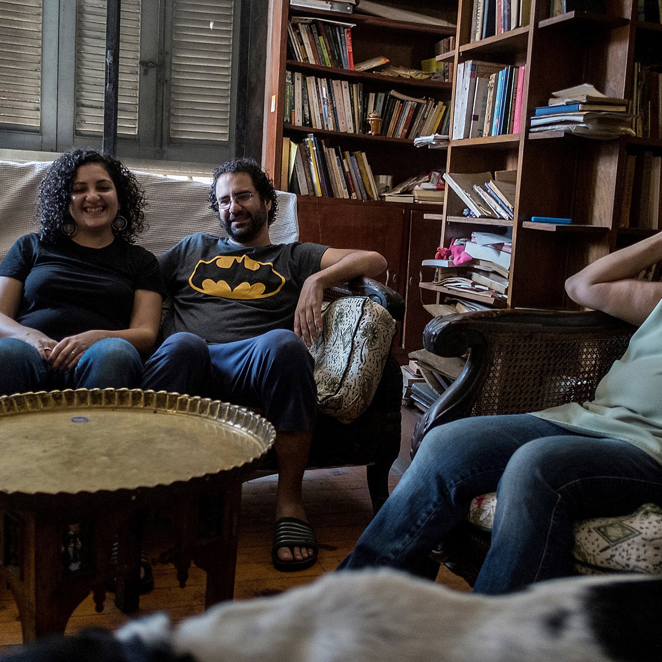 L'image montre un groupe de trois personnes assises dans un salon. Il y a deux femmes et un homme. La première femme, à gauche, sourit et porte un t-shirt noir. L'homme, au centre, porte un t-shirt avec le logo de Batman. La deuxième femme, à droite, a les cheveux bouclés et semble détendue. Ils sont assis sur des canapés, entourés d'étagères remplies de livres et d'objets décoratifs. Une ambiance conviviale et décontractée se dégage de la scène.